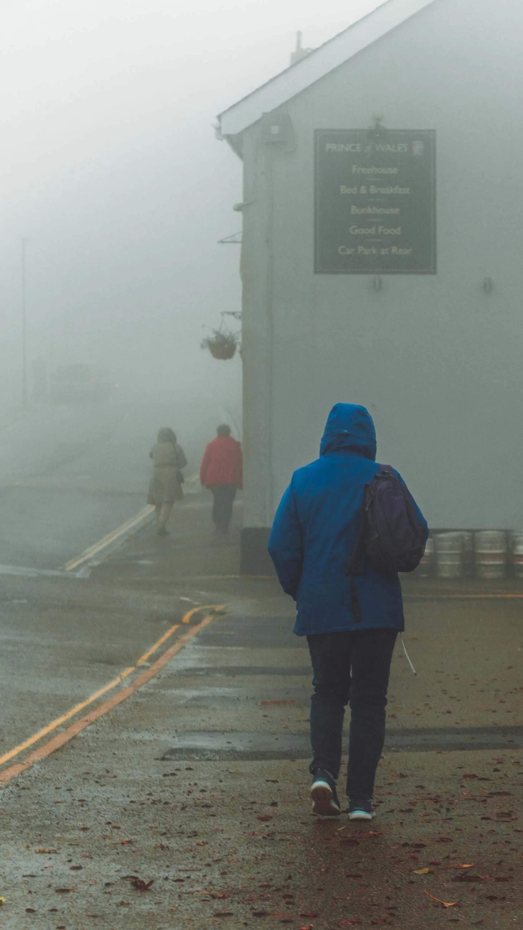 A person in a blue jacket and backpack walking on a foggy sidewalk with other pedestrians in the background near a building with a sign that reads "Prince of Wales" and lists amenities like a nightclub, restaurant, and car park.