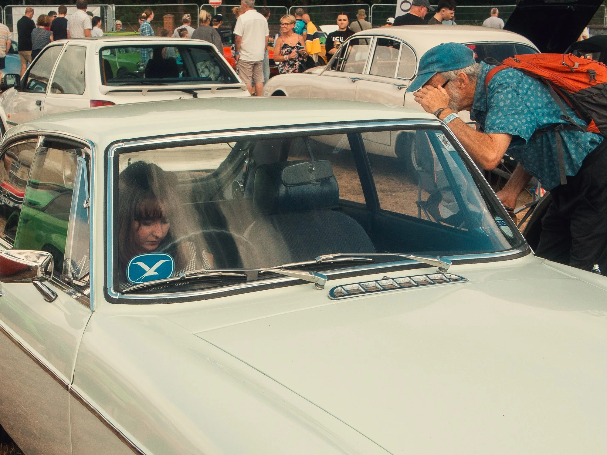 A man with a beard, wearing a blue cap and patterned shirt, leaning over a cream-colored vintage car during a car show, with a large crowd of people and other classic cars in the background.