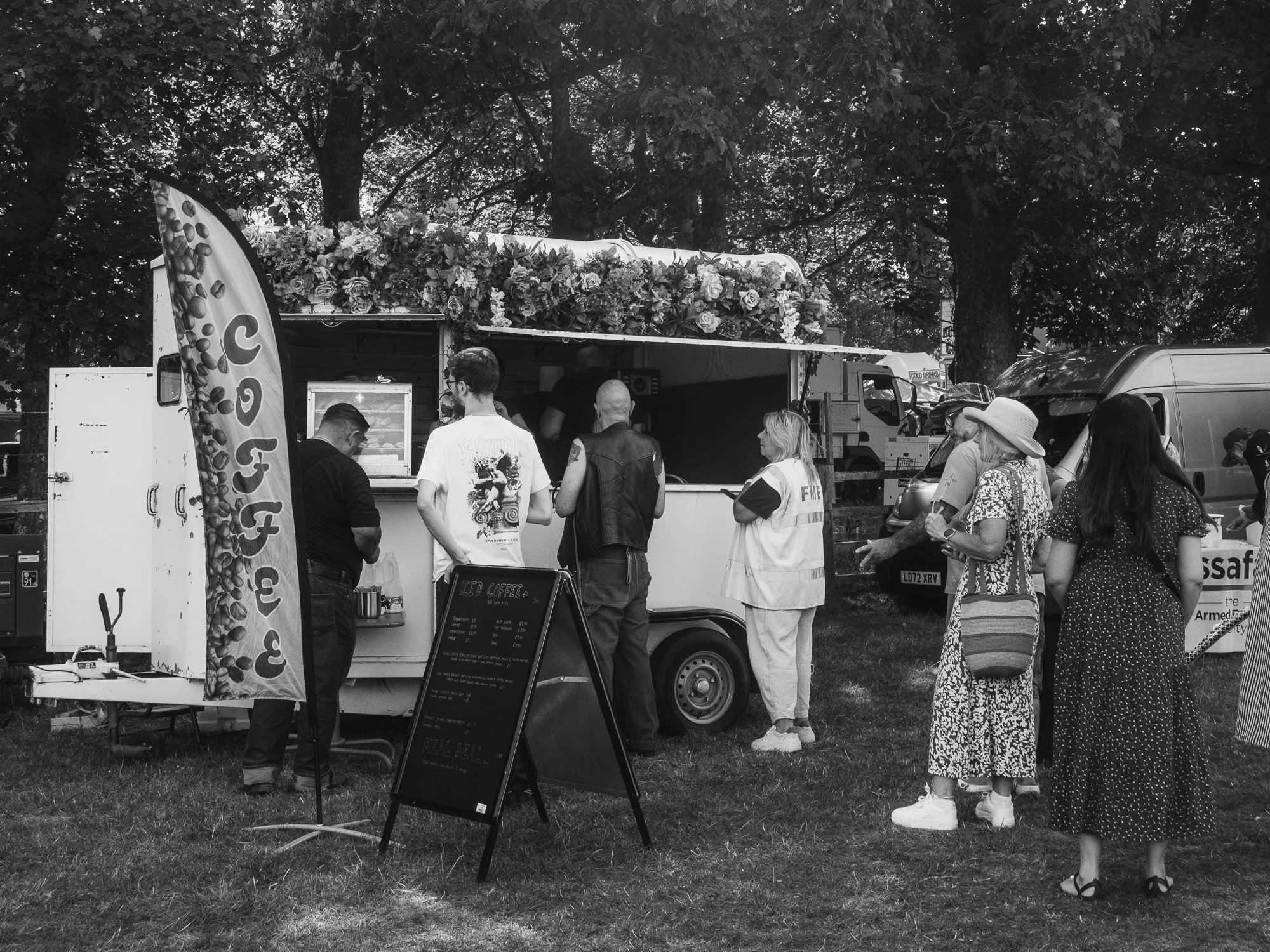 People standing in line at a food truck offering coffee at an outdoor event, with a large menu board and banners, surrounded by trees and parked vehicles.