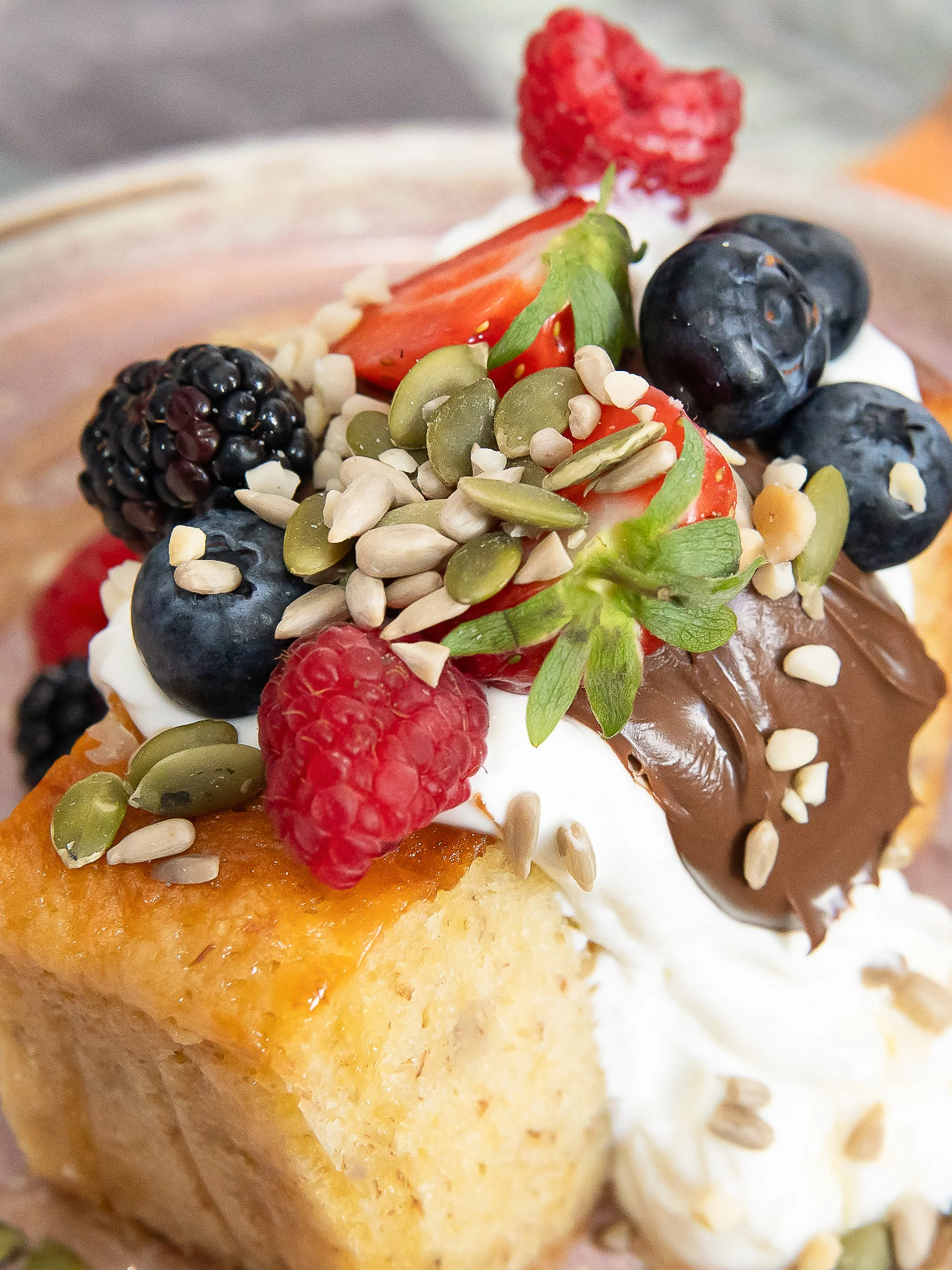 Close-up of a decorated dessert with fresh berries, sliced strawberries, blueberries, blackberries, raspberries, pumpkin seeds, sunflower seeds, and chopped nuts on whipped cream and cake.