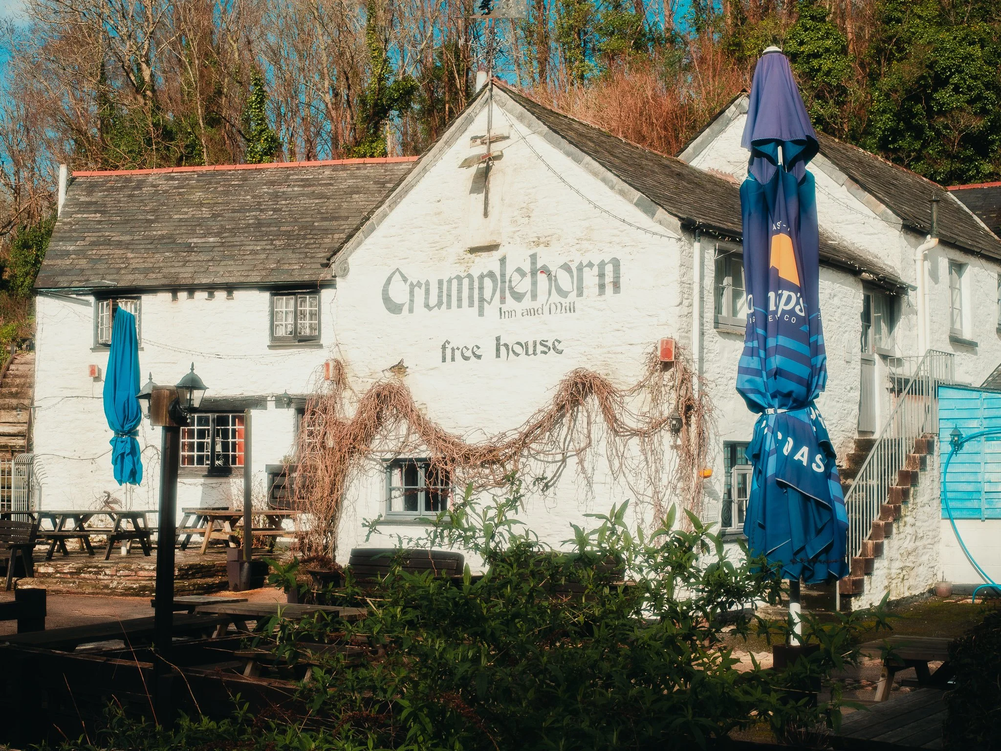 White building with sign that reads "Crumpthorn Inn and Mill free house" on the side, surrounded by trees and outdoor patio furniture with blue umbrellas.