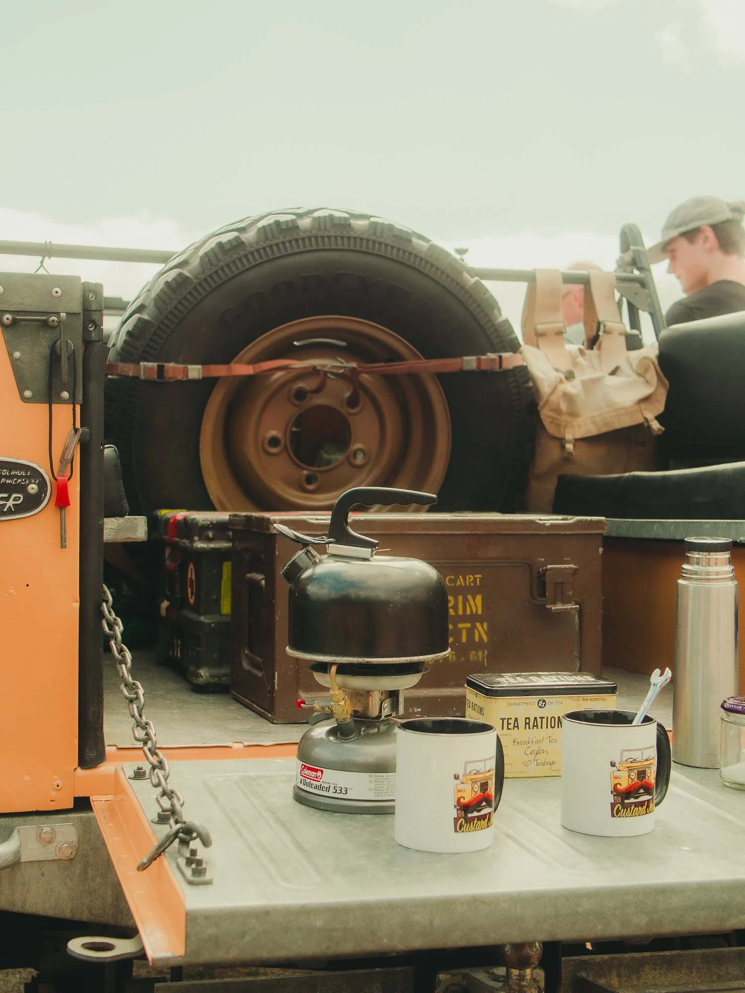 Three mugs with custard and tea branding are placed on a surface with a vintage tea ration container and other objects, with a background of a vehicle's cargo area containing spare tires and equipment.