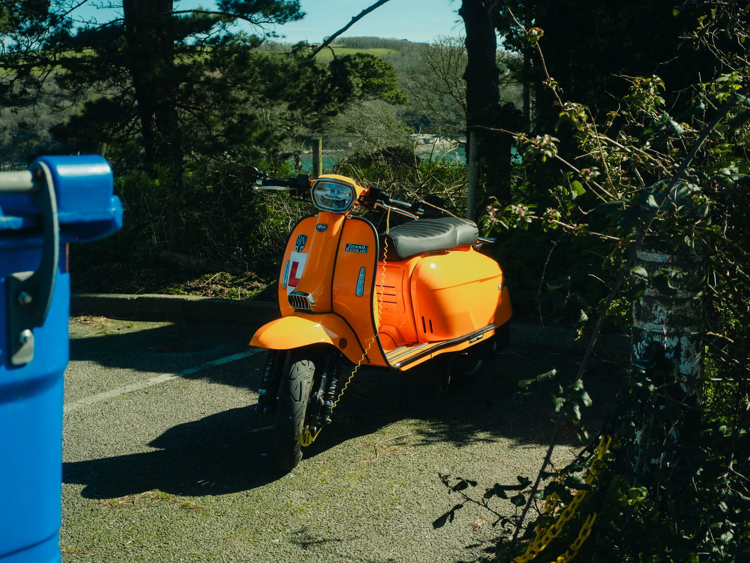 An orange scooter parked on a paved surface near a chain-link fence with green plants. In the background, there are trees, hills, and a blue sky.