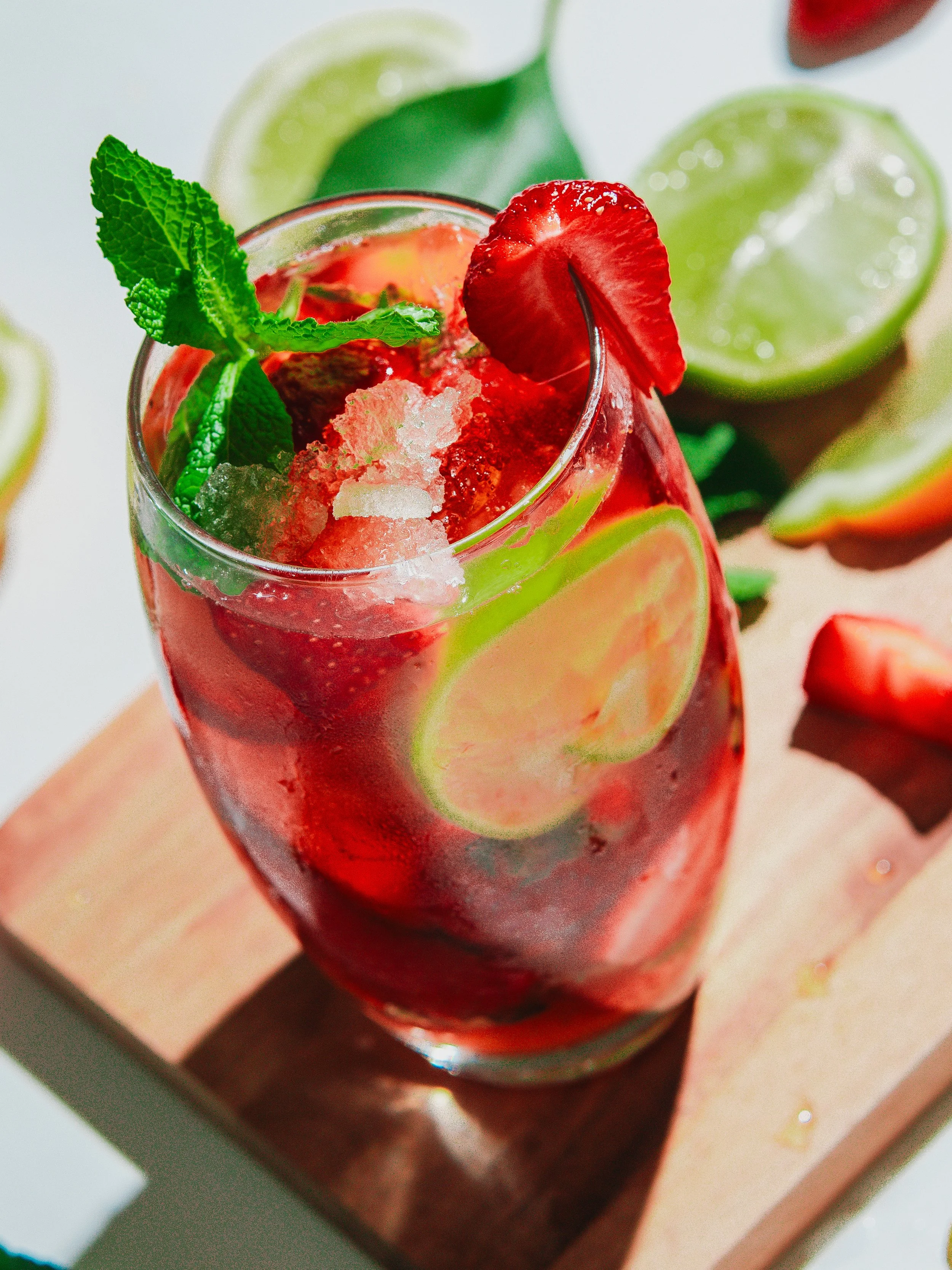 Glass of strawberry-lime mocktail garnished with a strawberry slice, mint, and ice, with lime and strawberry slices around it.