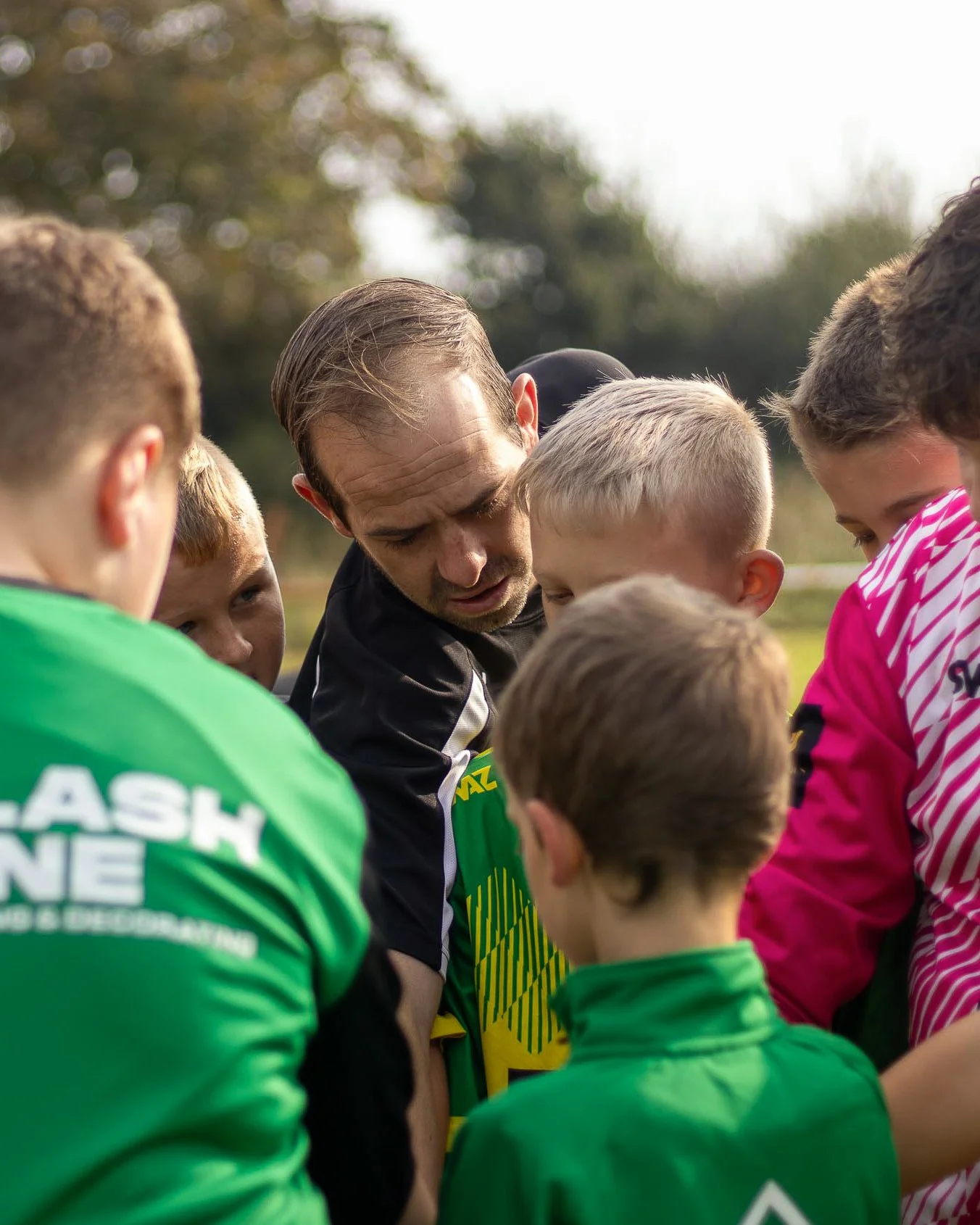 A man, likely a coach or team leader, surrounded by young children in sports jerseys on a field, all appearing to be listening intently and huddled together.