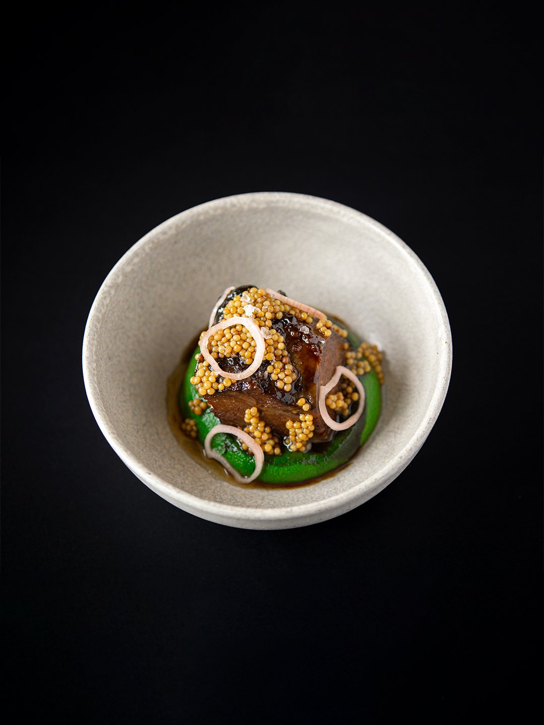 A bowl of gourmet Asian cuisine with braised beef, mustard seeds, sliced green onions, and a dark sauce, served in a white ceramic bowl against a black background.