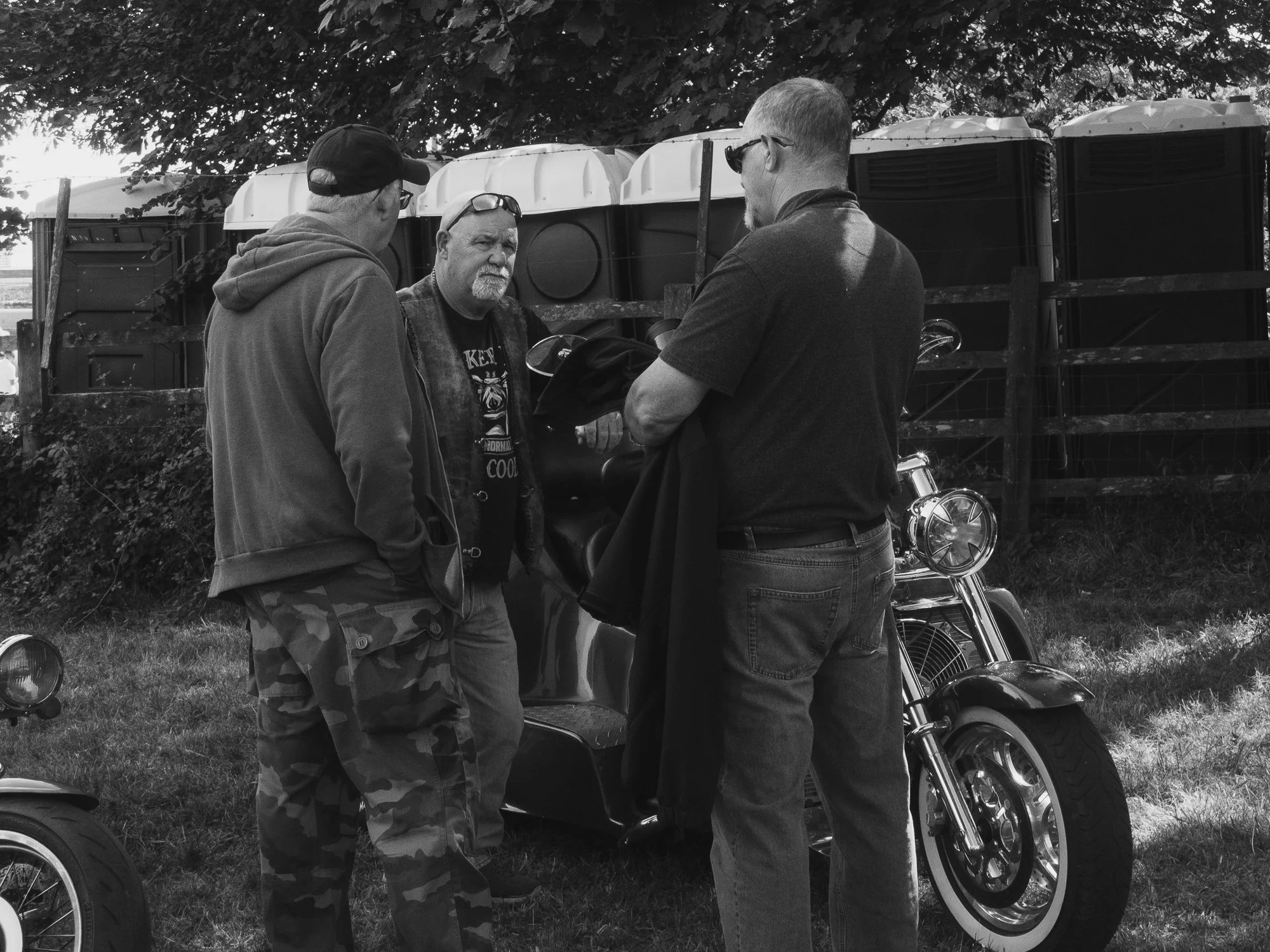 Three men stand beside a motorcycle and a golf cart outdoors, having a conversation. One man wears a hoodie and cap, another wears a leather jacket and sunglasses, and the third has a beard and glasses. A fence, trees, and some storage containers are