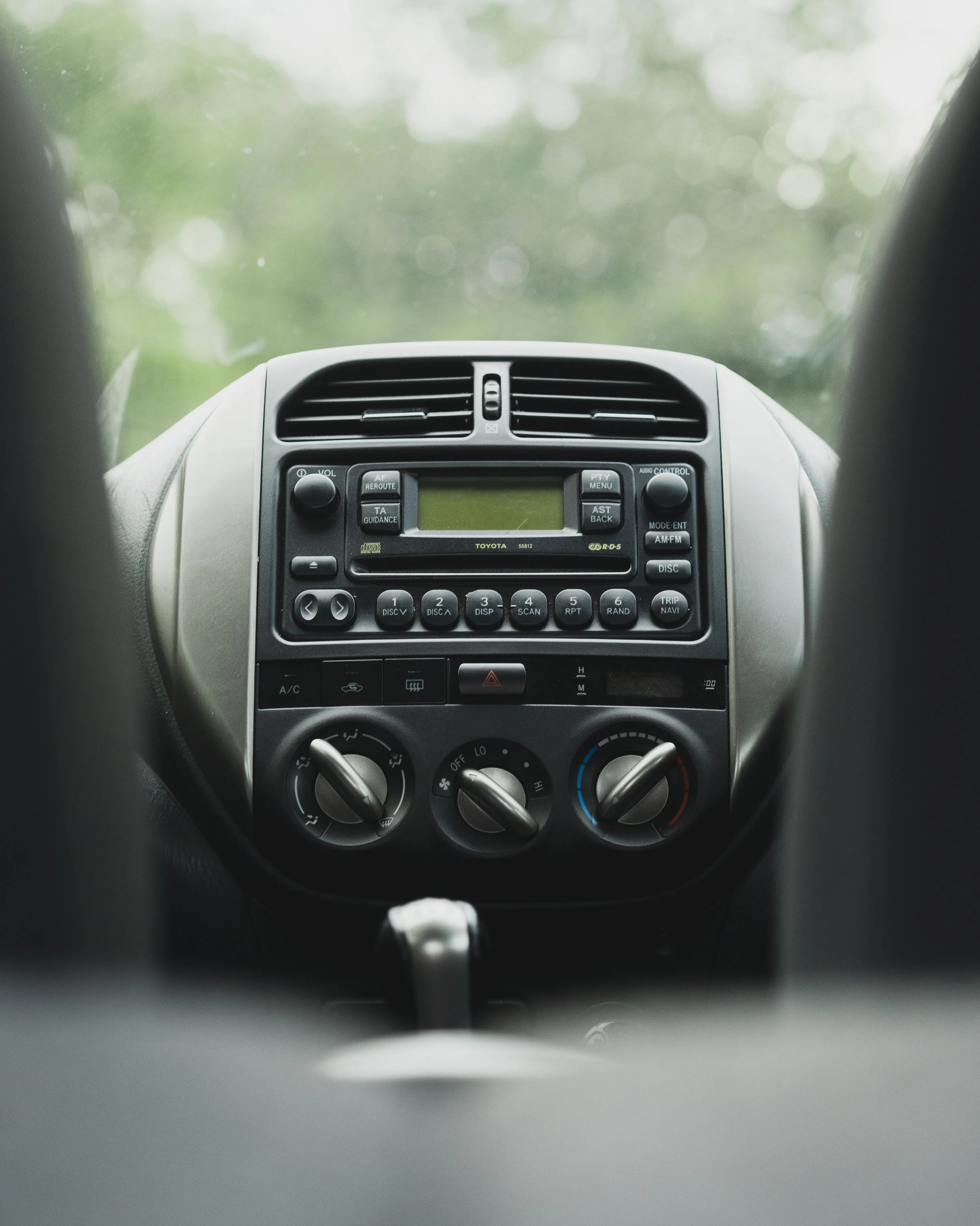 Front dashboard of a car with radio, air conditioning, and control knobs, seen from the driver's perspective with a blurred view of outside through the windshield.