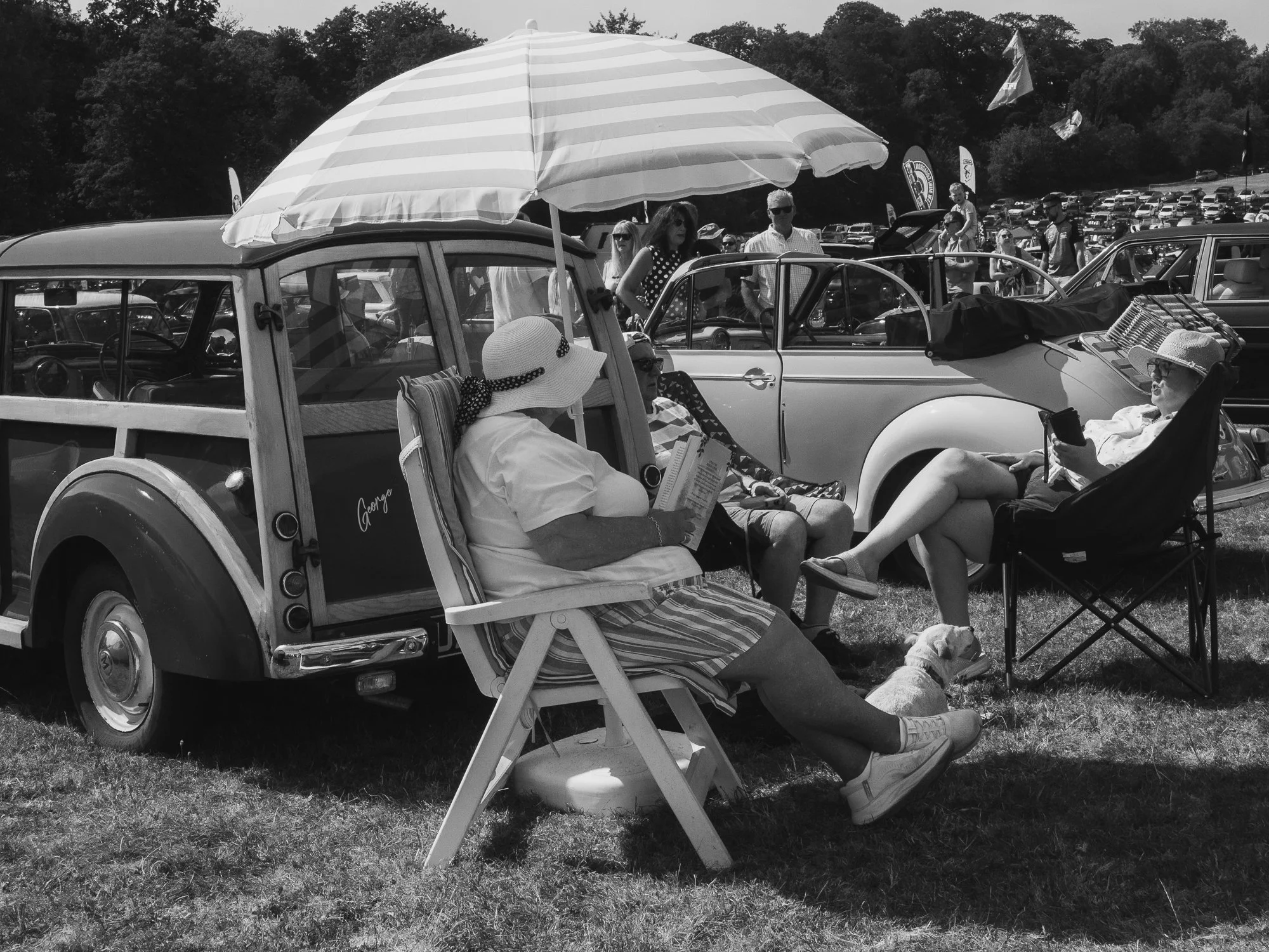 People relaxing at an outdoor car show with vintage cars and tents in the background.