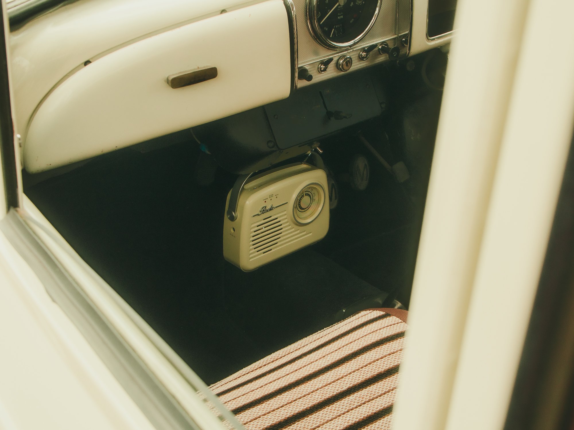Inside view of a vintage car dashboard and floor, featuring an analog clock, radio, pedals, and a glove compartment, with a striped floor mat.