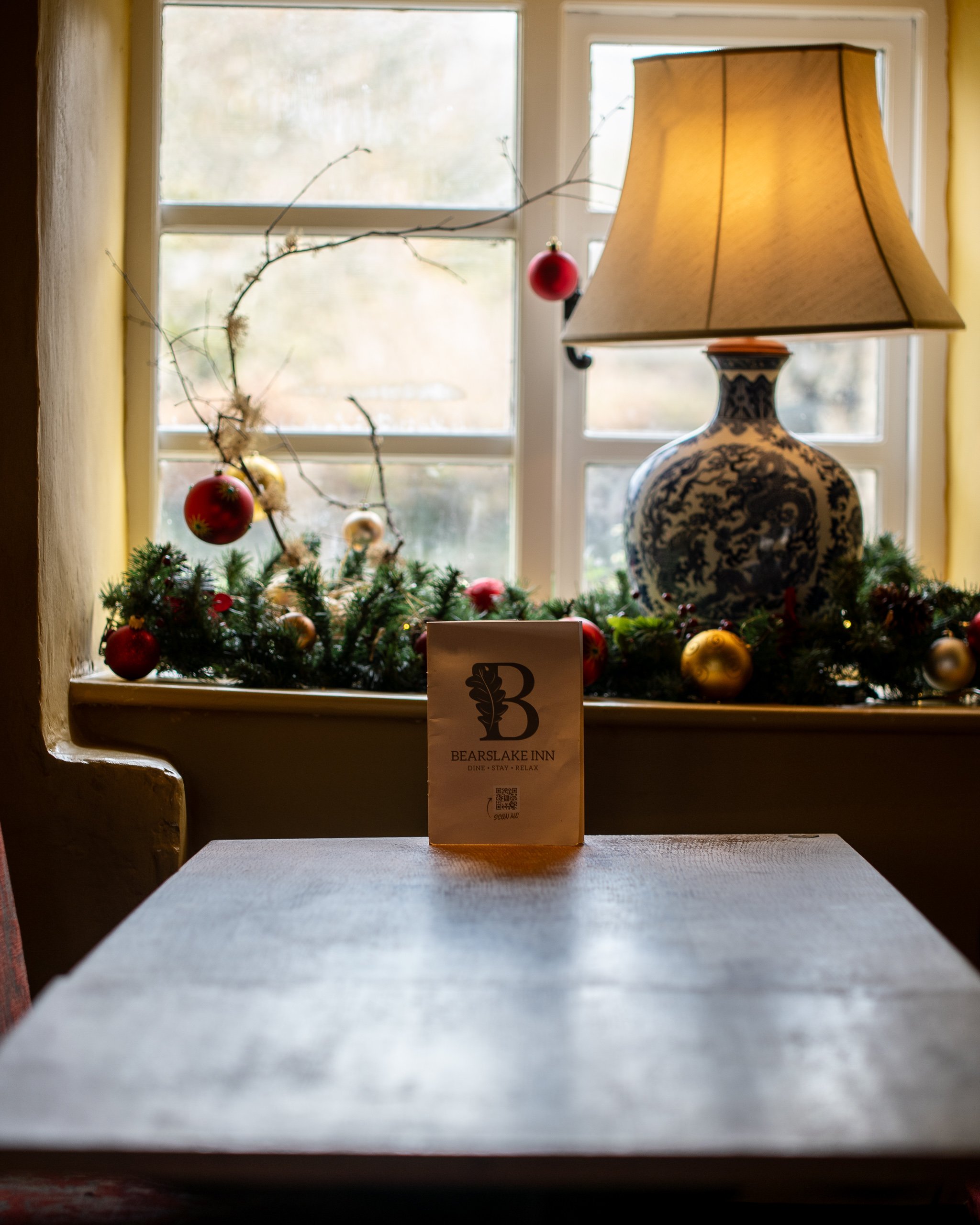 A wooden table in front of a window decorated with a Christmas garland, red and gold ornaments, and a blue and white porcelain lamp, with a menu from Bearsake Inn on the table.