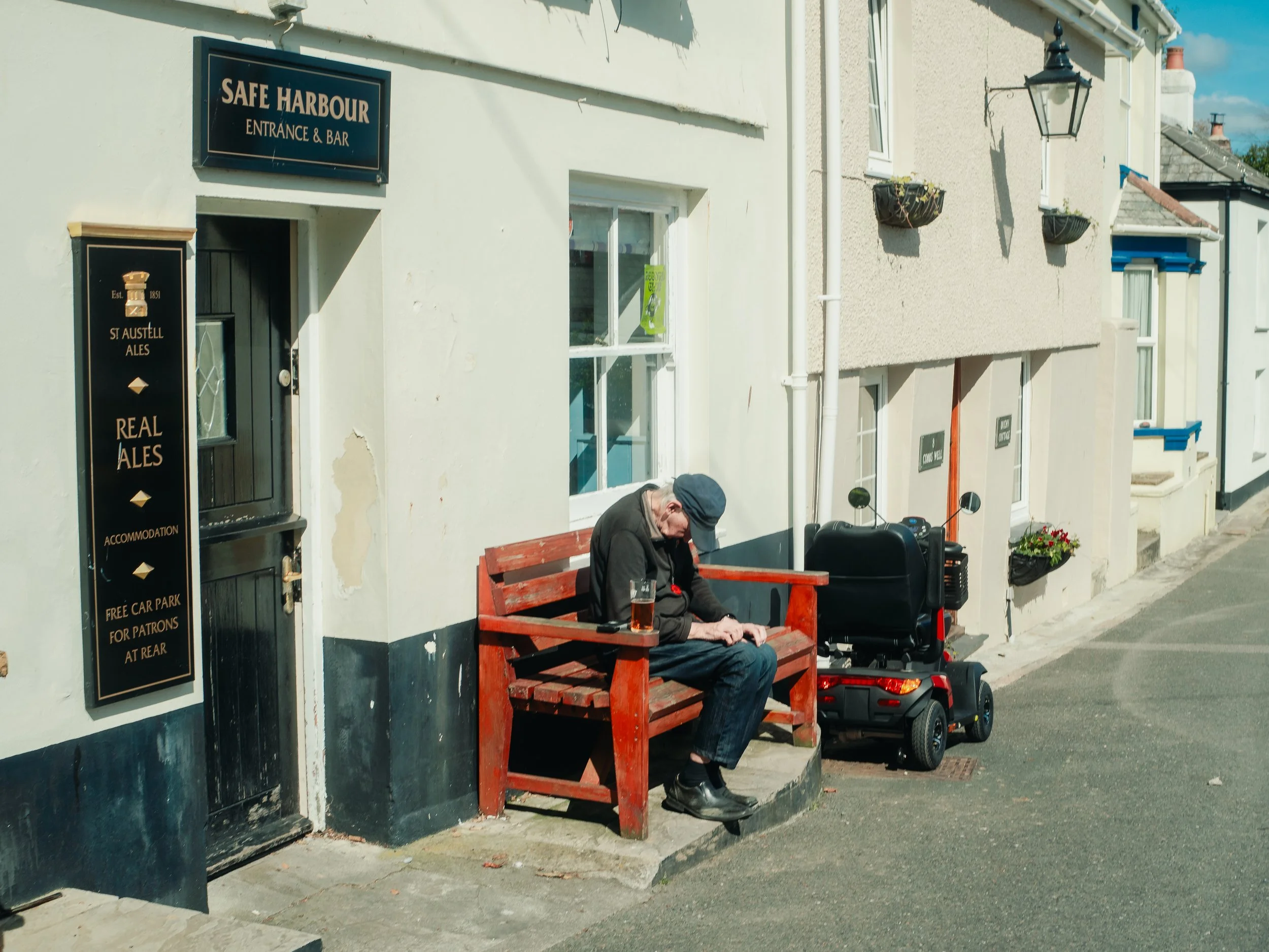 A man wearing a cap sitting on a red wooden bench on a sidewalk outside a building, with his head down. There is a pint of beer on the bench beside him and a motorized scooter nearby. The building has signs for a bar and accommodation, and there are 