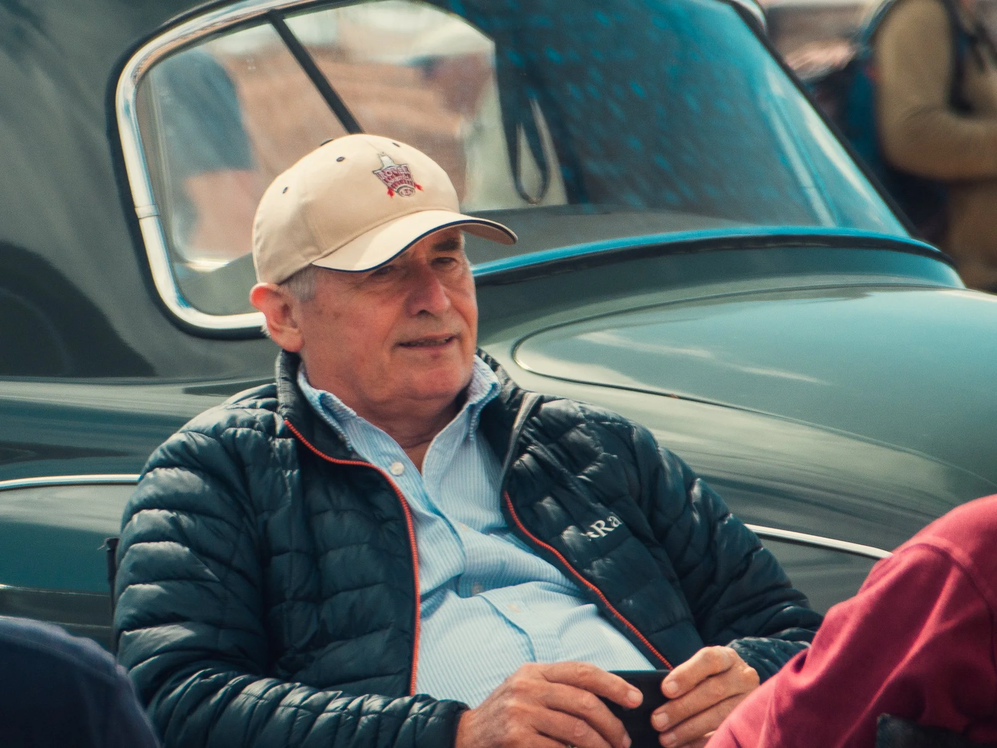 A man is sitting in a classic car, wearing a beige baseball cap with a logo and a black quilted jacket. The car has a blue interior, and the man appears to be looking at his hands or an object he is holding.