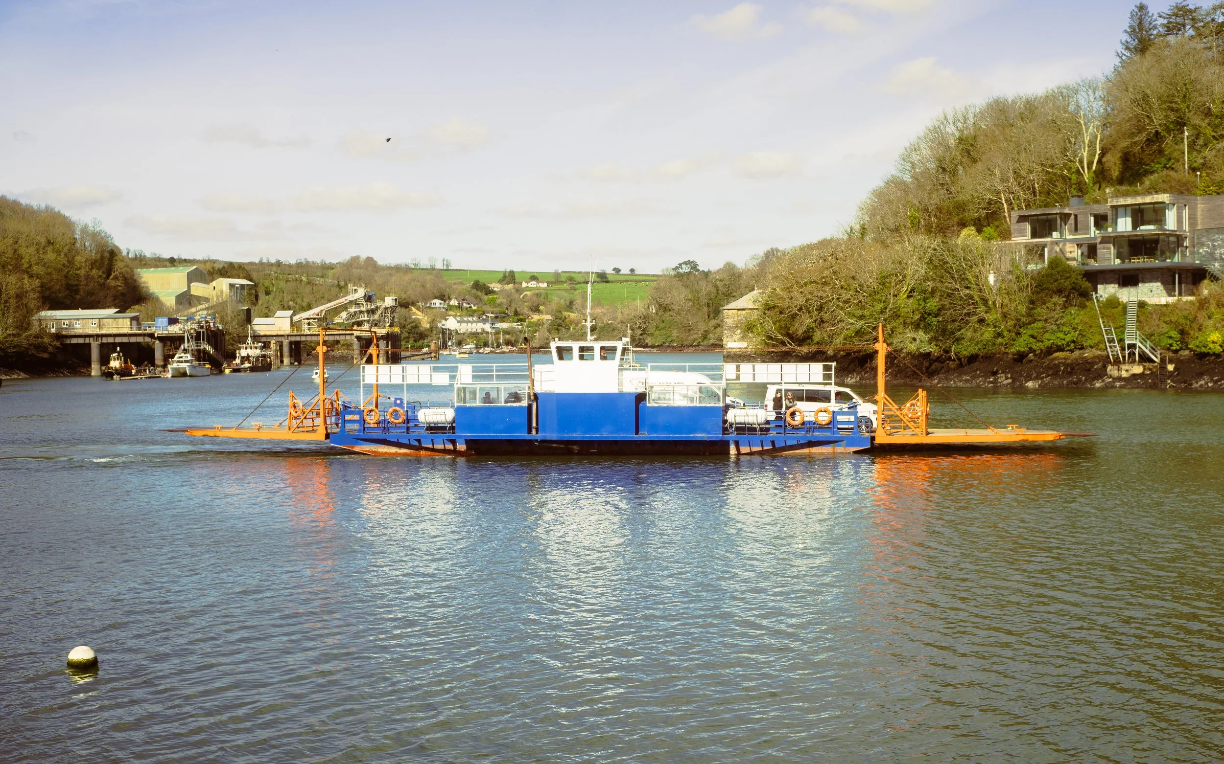 A blue ferry boat on a river with houses, trees, and hills in the background under a partly cloudy sky.