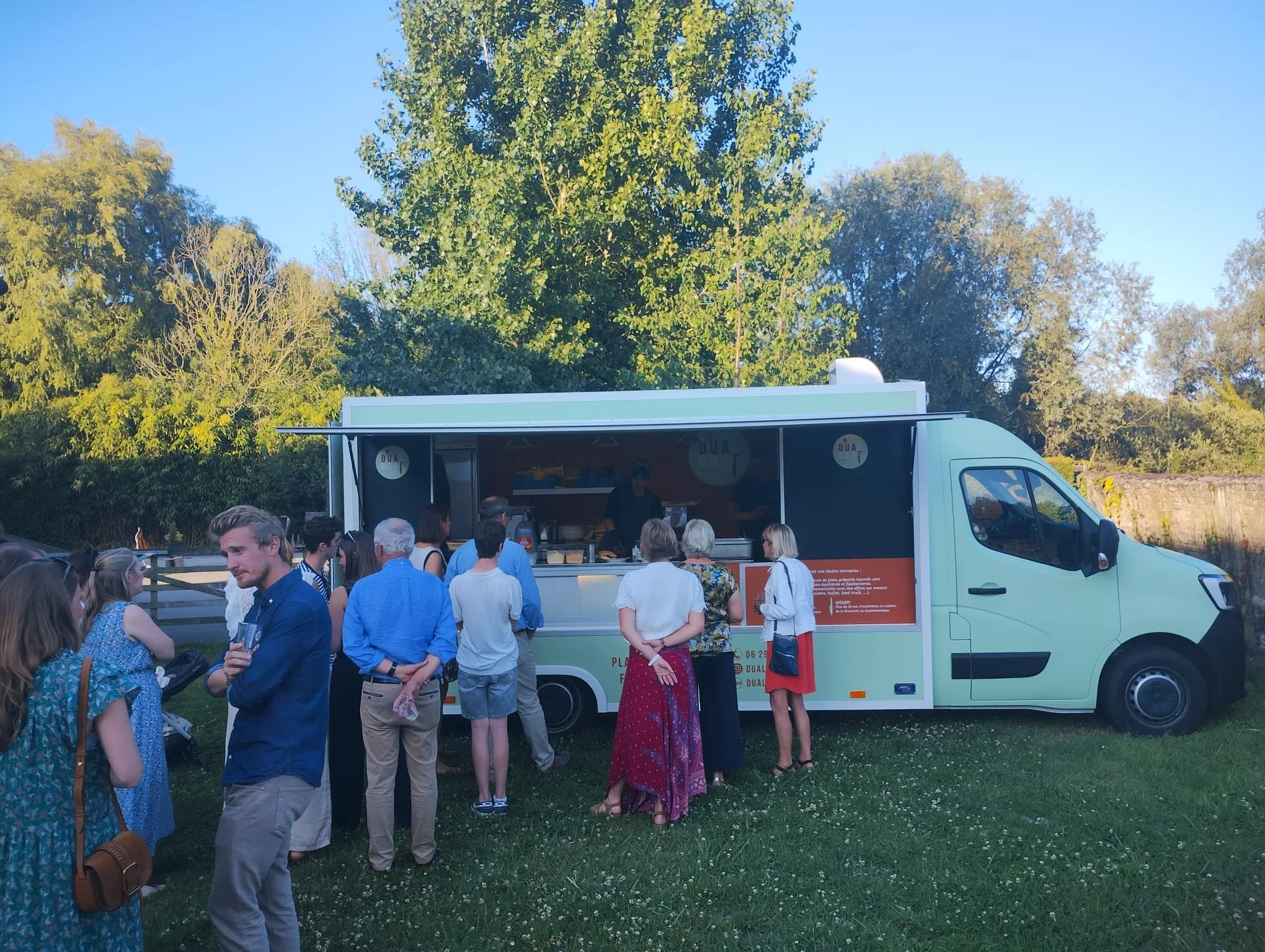 Une file de personnes faisant la queue devant le food truck "Dual Food" dans un parc avec des arbres en arrière-plan par une journée ensoleillée.