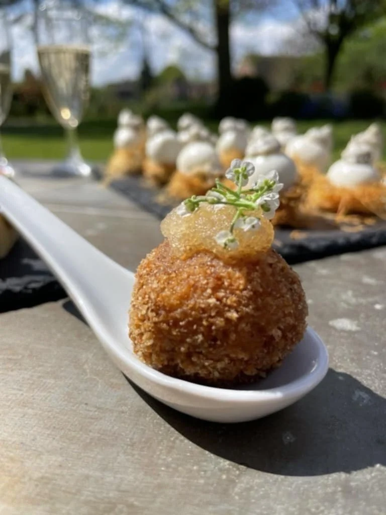 Boulettes de risotto frites garnies de fleurs comestibles sur une cuillère en porcelaine, avec une présentation de bouchées similaires en arrière-plan en plein air.
