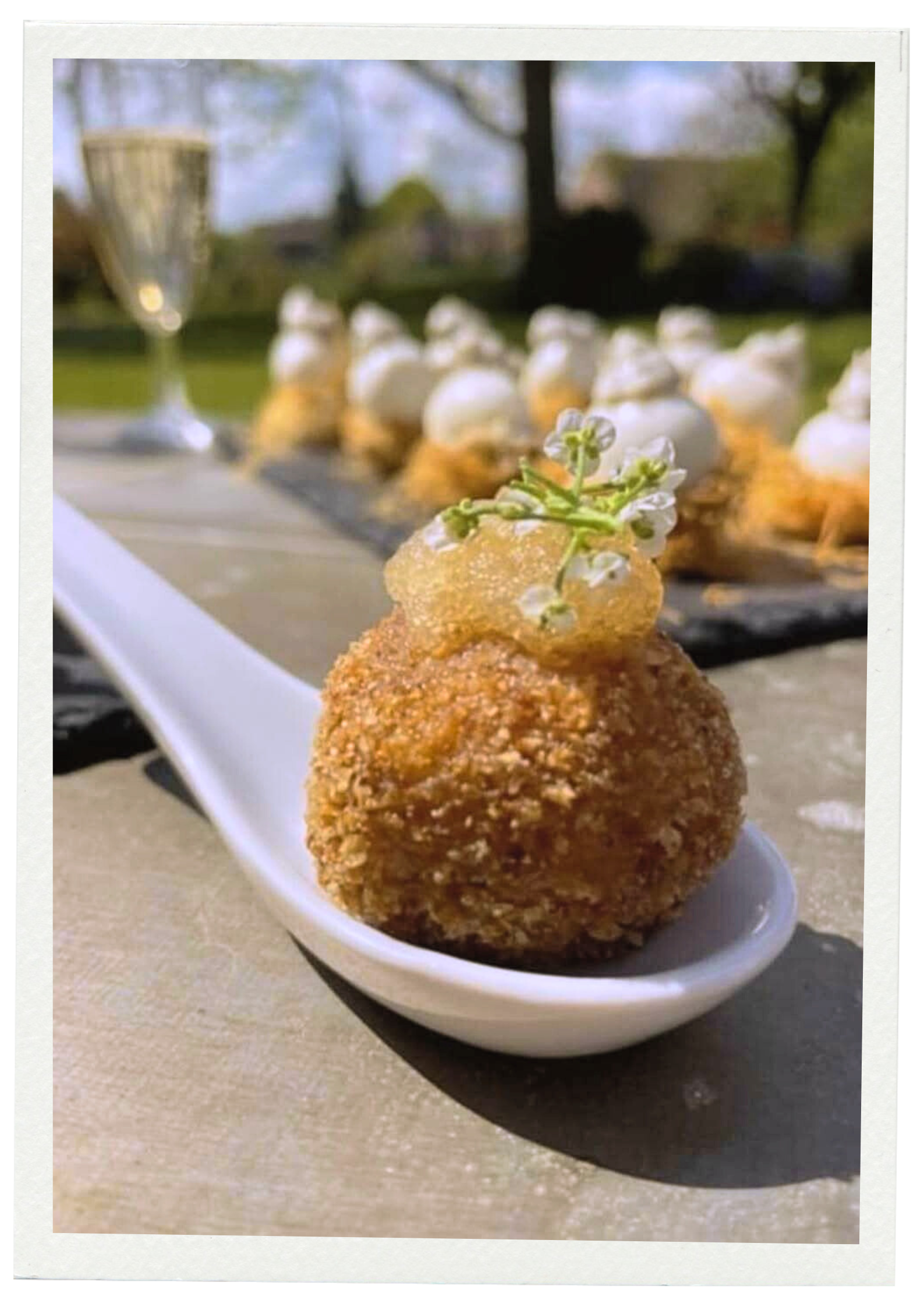 Une bouchée de nourriture frites garnie d'une petite fleur blanche sur une cuillère blanche, avec d'autres bouchées en arrière-plan et un verre à champagne devant un fond de parc en plein air.