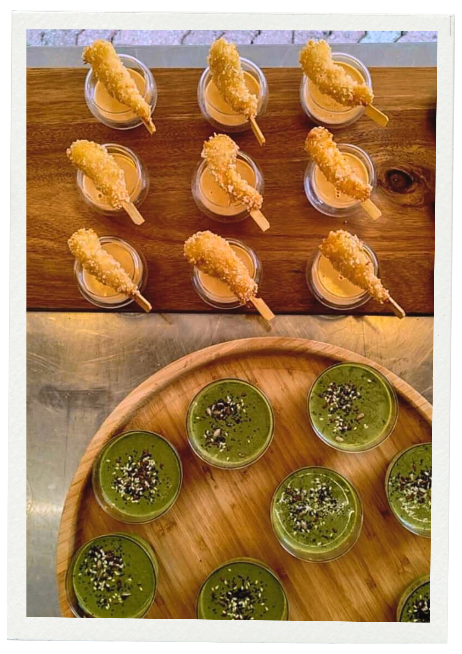 Gobelets contenant des desserts ou boissons vertes garnis de graines, disposés sur une assiette en bois, et une planche en bois avec des petits fours ou aliments frits, chacun avec une brochette, posés sur une surface en métal.
