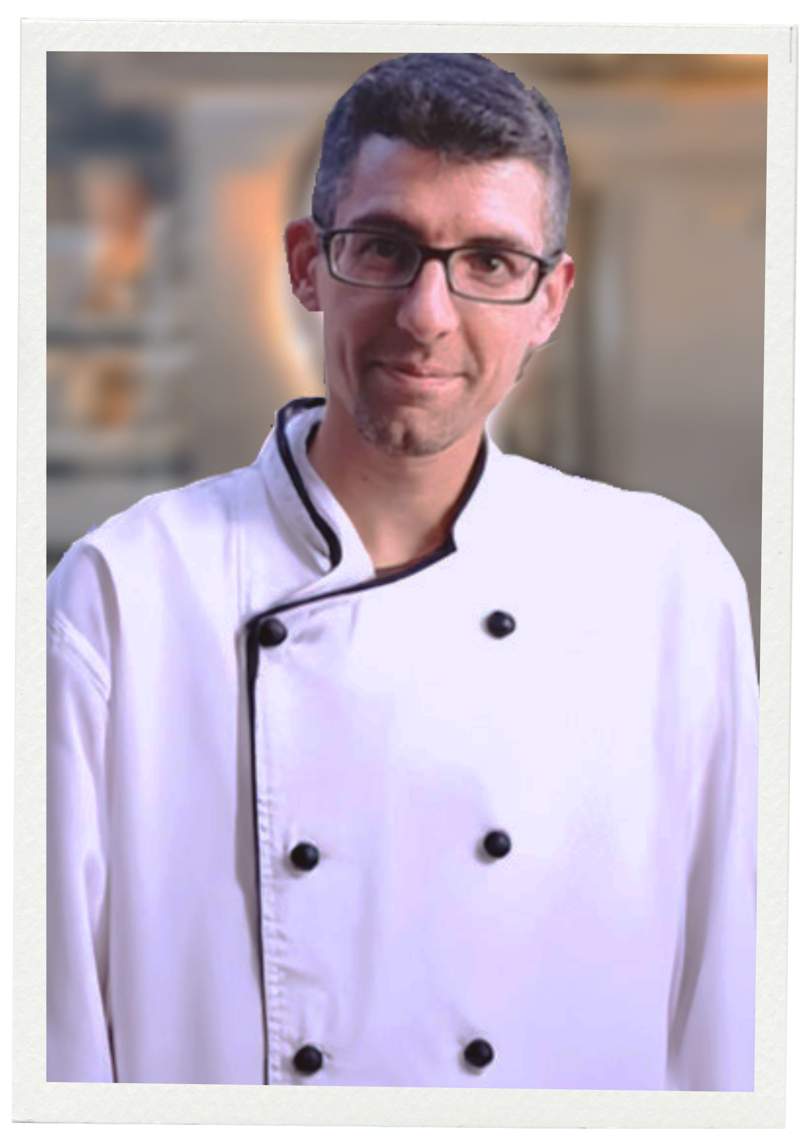 Gregory en blouse de chef cuisinier, portant des lunettes, dans une cuisine.