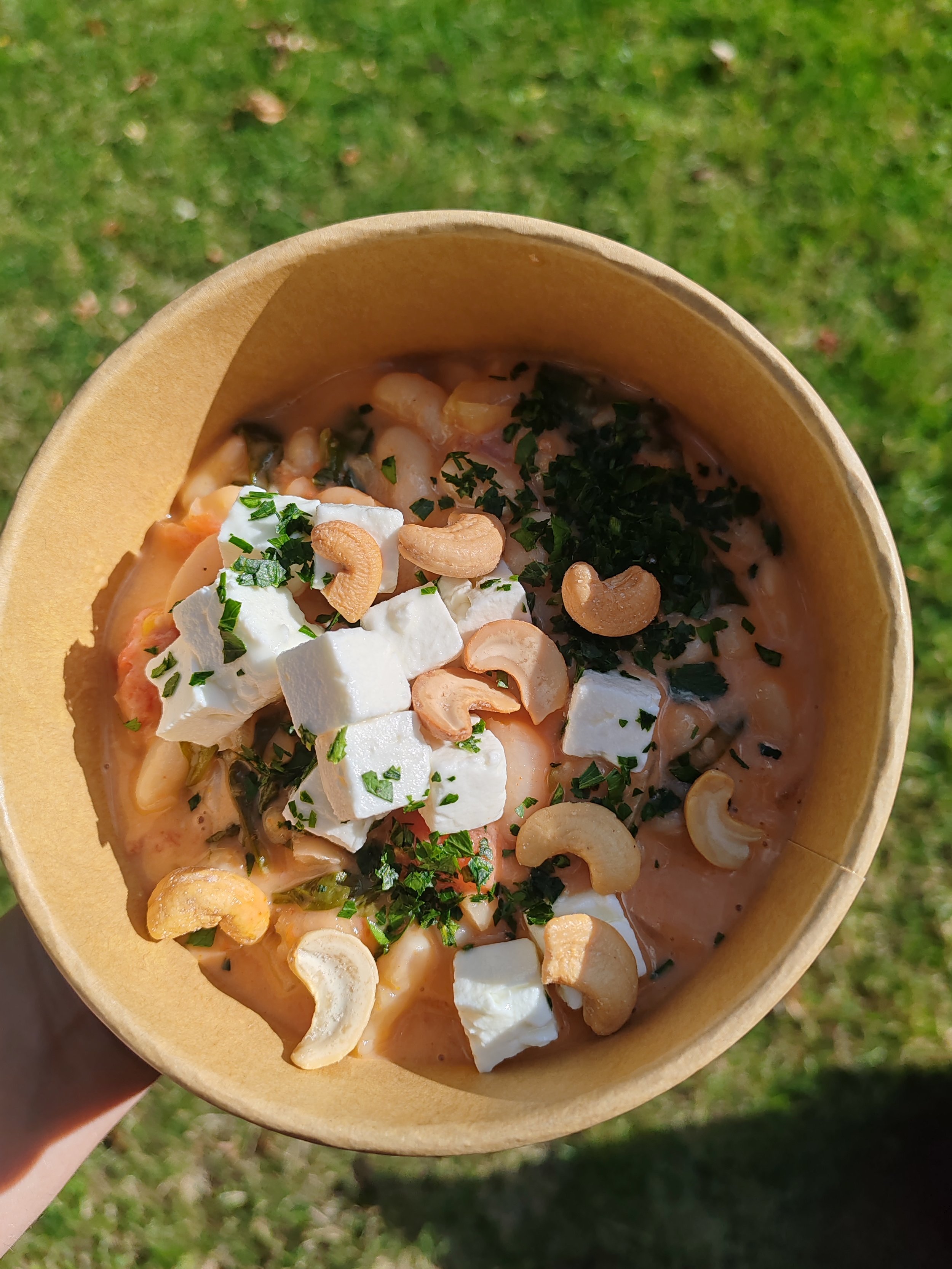 Une coupe en papier contenant un plat de fruits de mer avec du fromage, des noix de cajou, et des herbes, vue de dessus, avec un fond d'herbe verte.