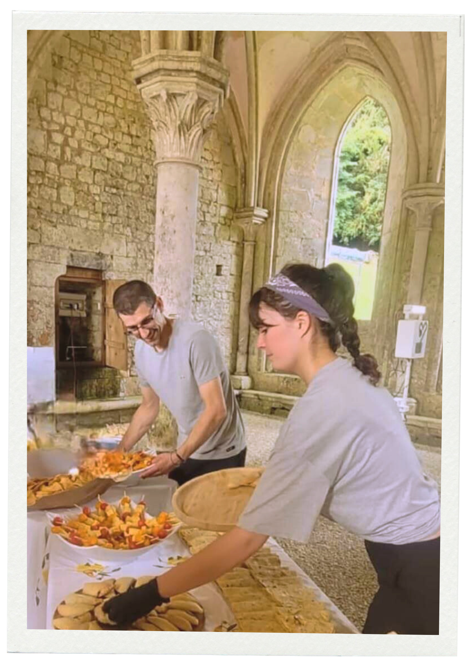 Bertille & Gregory servent des plats et des desserts sur une table dans un lieu ancien avec des arches en pierre.