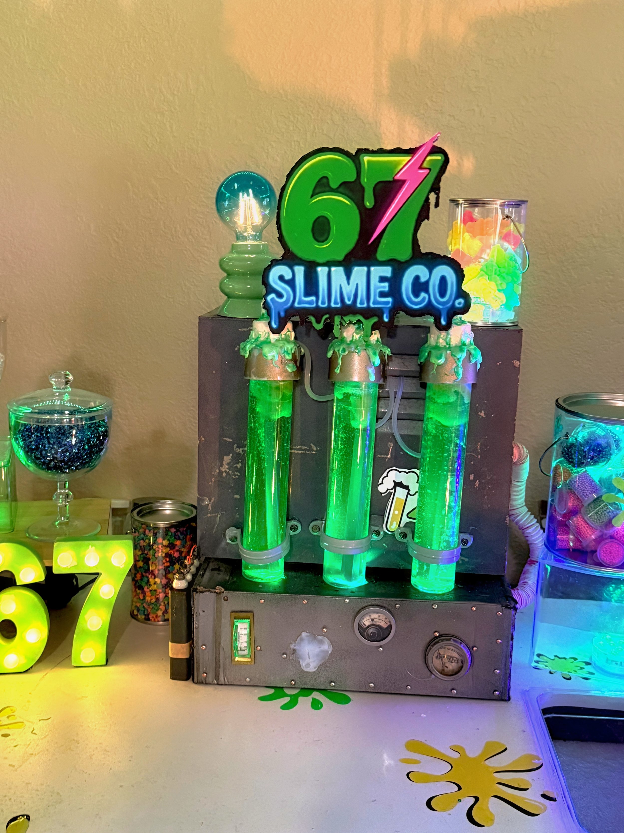 Colorful slime machine with three glowing green tubes, a neon sign reading '67 Slime Co.', and various containers of slime and decorations on a decorated table with yellow and black splashes.