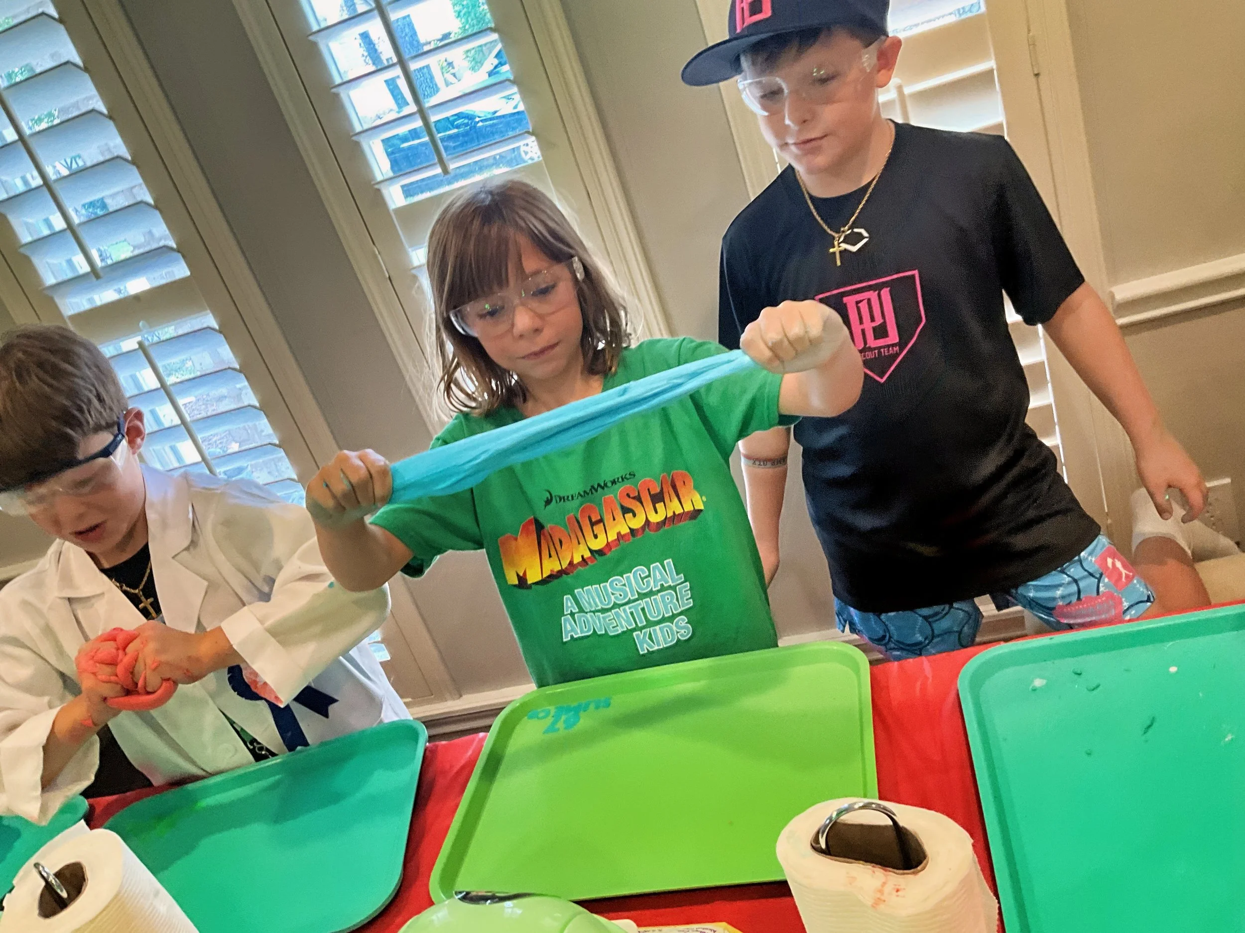 Children engaging in a hands-on science activity, hosted by 67 SLIME Co.  at a 67 mobile lab, with colorful containers, wearing safety goggles, in a well-lit room with windows and closed plantation shutters.