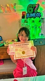 Girl in pink hoodie holding a certificate and smiling in a room with slime-themed decorations