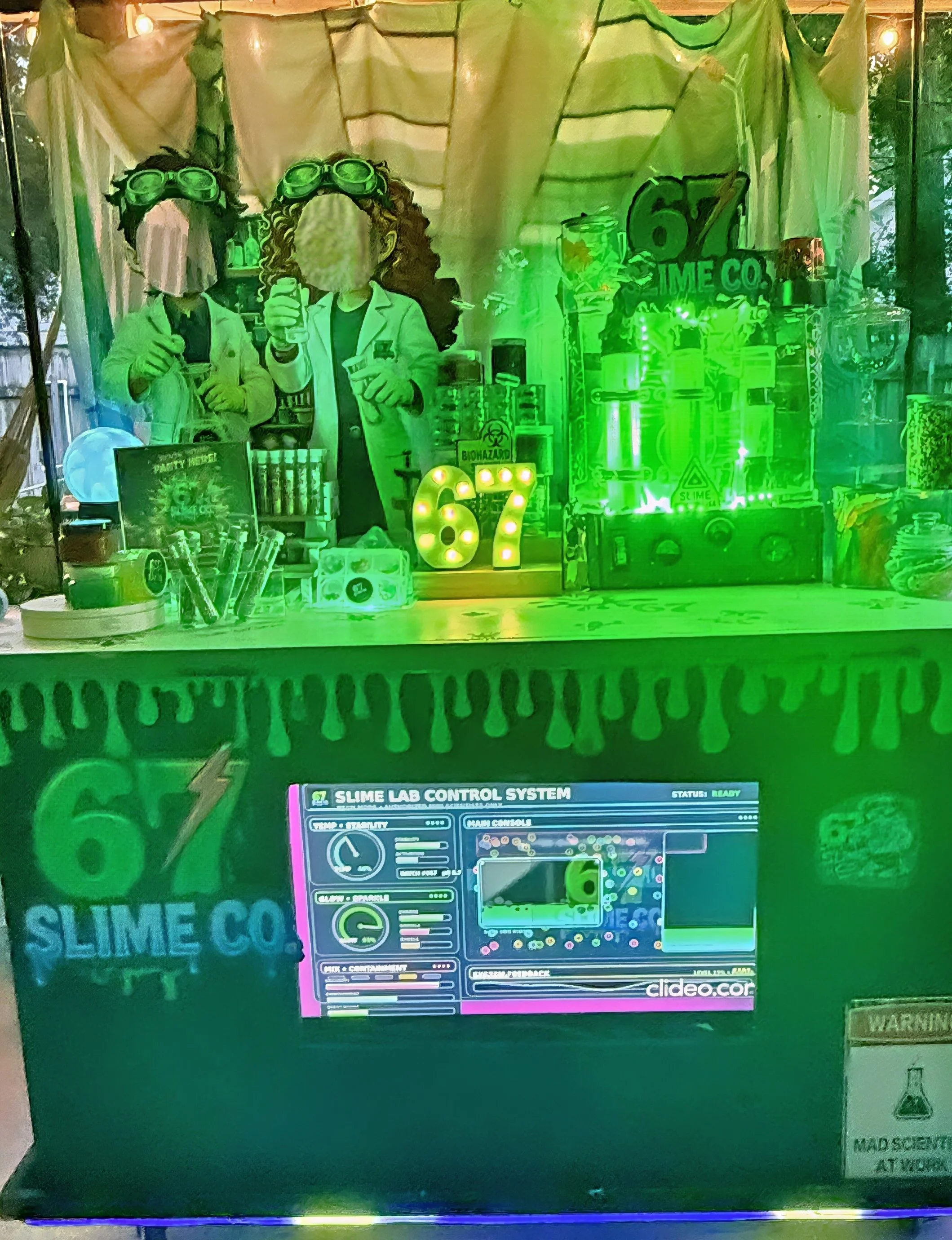 A science-themed display featuring two cutouts of scientists in white lab coats, wearing goggles, with one giving a thumbs up and the other holding a test tube. To the right, there are scientific equipment and a sign for 'Slime Lab Control System' with colorful graphics. The background includes a large number '67' and various scientific icons, and the scene is illuminated with green lighting.