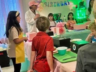 Children at a birthday party or celebration, with a woman serving or preparing food at a decorated table with pink and green accents.