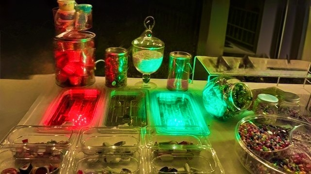 Assorted glass jars and containers filled with colorful beads, buttons, or craft supplies, illuminated by red and green lights make up the 67 Slime Co. Mobile Lab.  Houston's Mobile Slime Lab Birthday Experience.