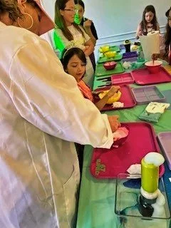 Kids at a science or activity table with trays, supplies, and a person demonstrating or guiding.