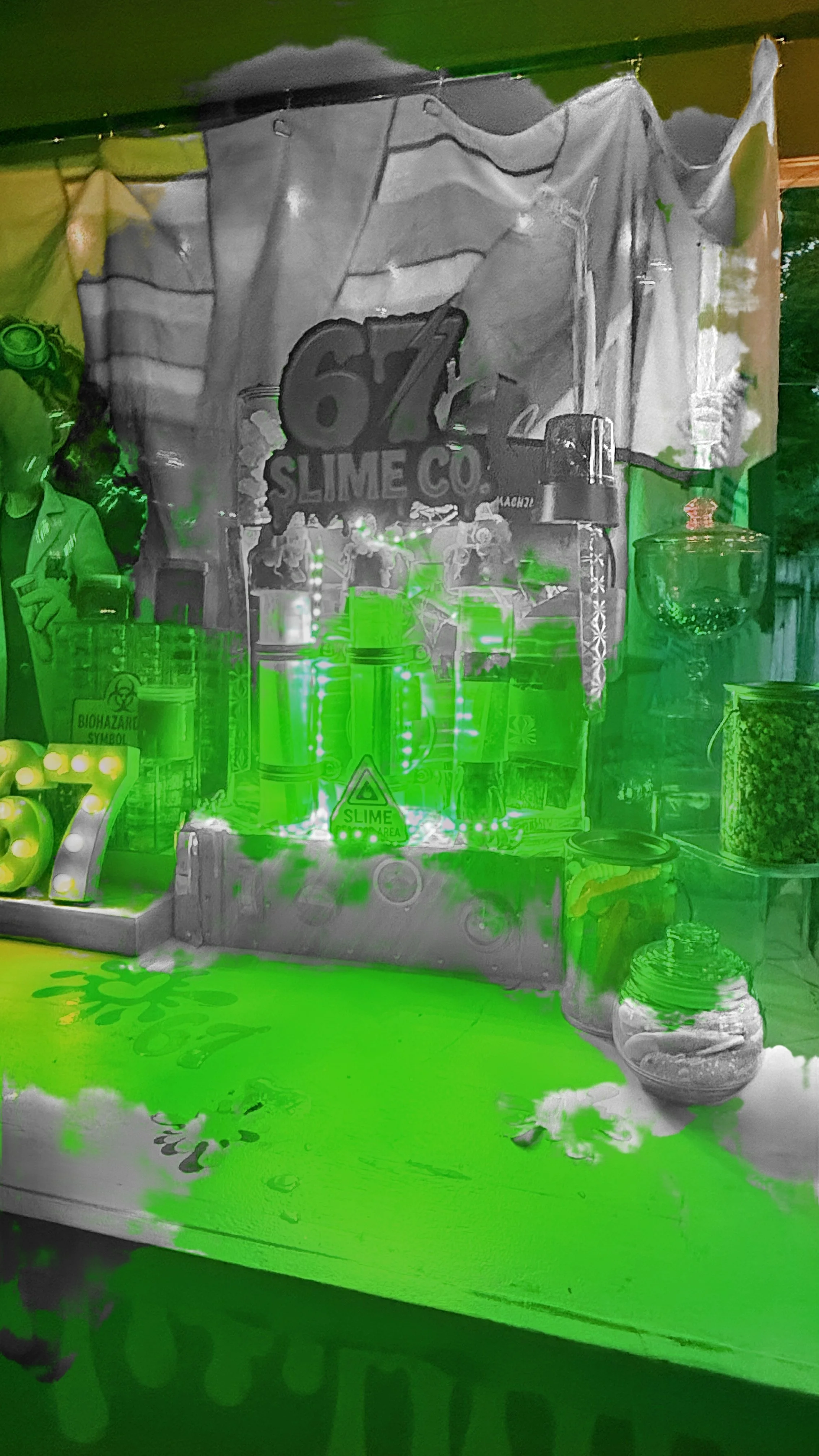 A display booth of slime from '67 Slime Co.' with various slime containers, decorations, and signs, illuminated with green lighting.