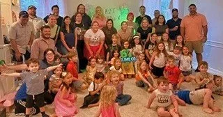 A large group of children and adults posing together in a colorful room with a bright green screen and decorations.