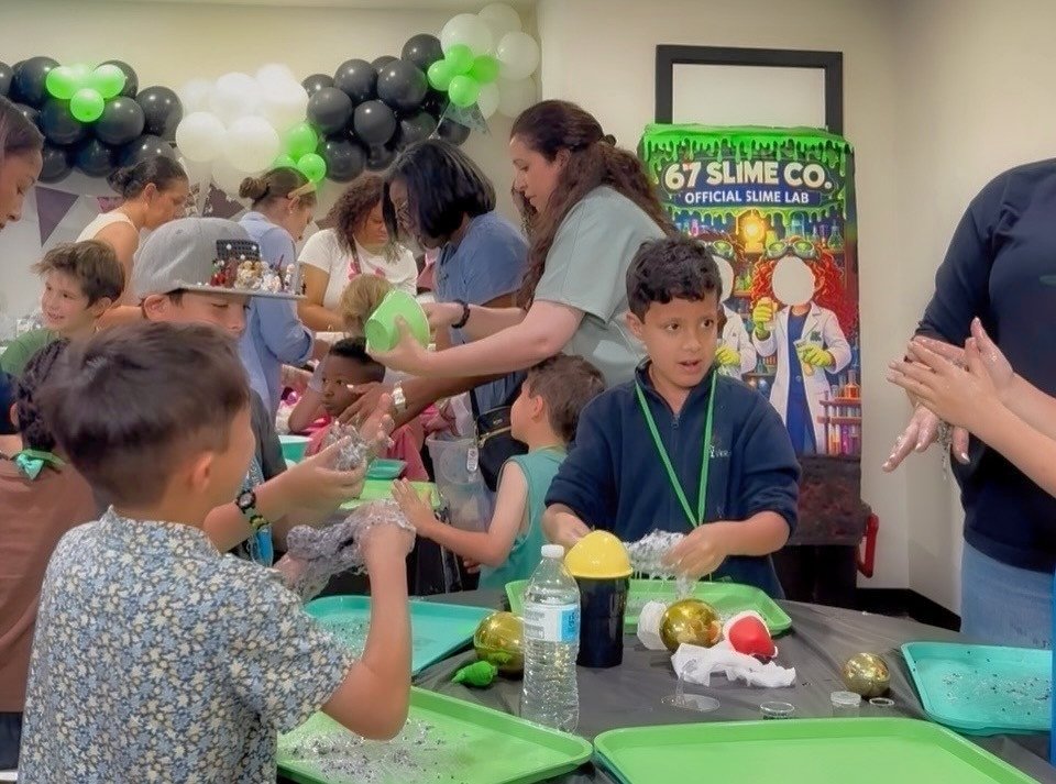 Children and adults at a slime-making party with green and gold decorations, working with slime and slime-making supplies.