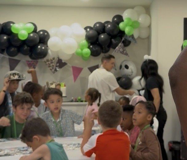 Children at a birthday party decorated with black, white, and green balloons, a panda face balloon, and colorful pennant banners.