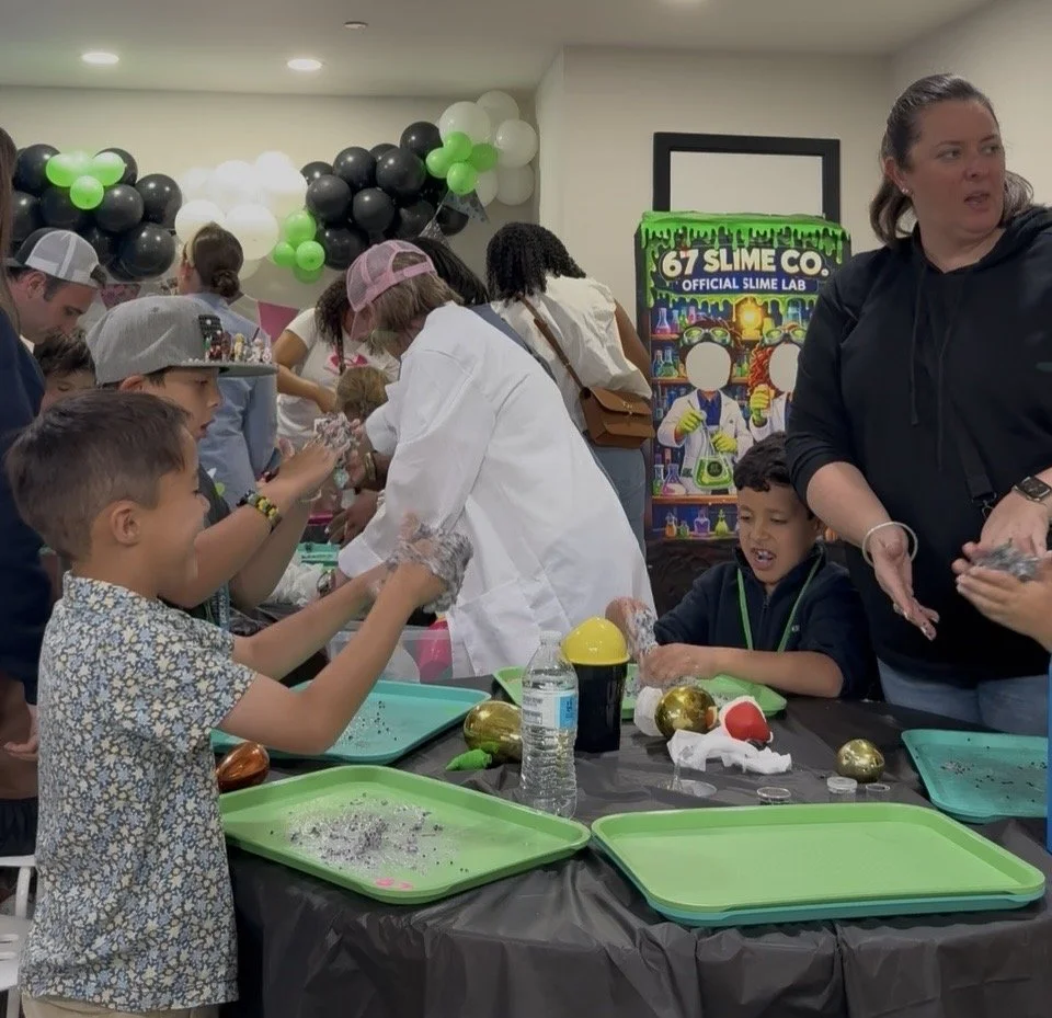 Children and adults at a slime-making event, with a table full of slime supplies and a slime company sign in the background.