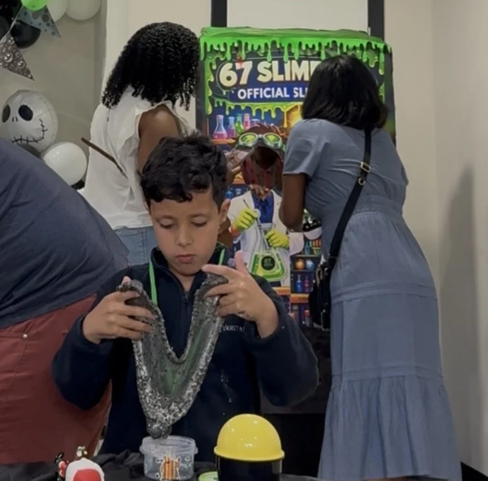 A young boy holding his custom slime he had created. at a Birthday party with a '67 Slim' slime activity station in the background. Other children and adults are engaging in activities.