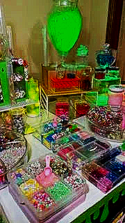 A colorful display of nail art supplies, including various nail polish bottles, nail gems, and decorations arranged on a table with a green heart-shaped balloon in the background.