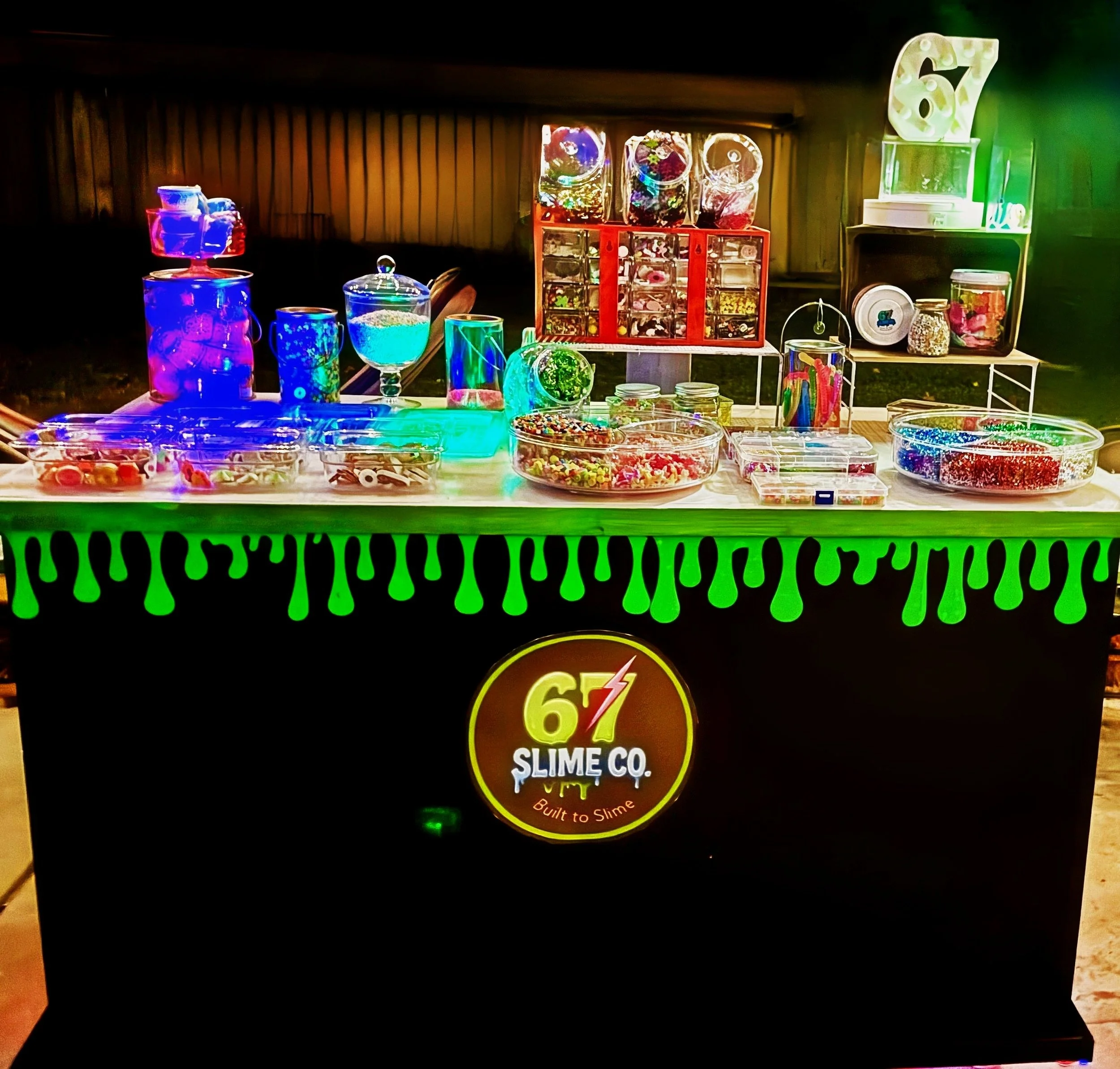 A slime-making kiosk with colorful slime containers and decorations. The kiosk has a black front with green slime drips and a logo that reads "67 Slime Co." It displays various slime supplies and accessories on the table and shelves.