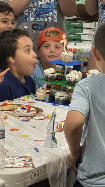 A birthday party with children gathered around a table decorated with balloons and confetti. A boy wearing a red cap is looking at a table with cupcakes and a colorful birthday cake with a candle. Other children are celebrating and smiling.