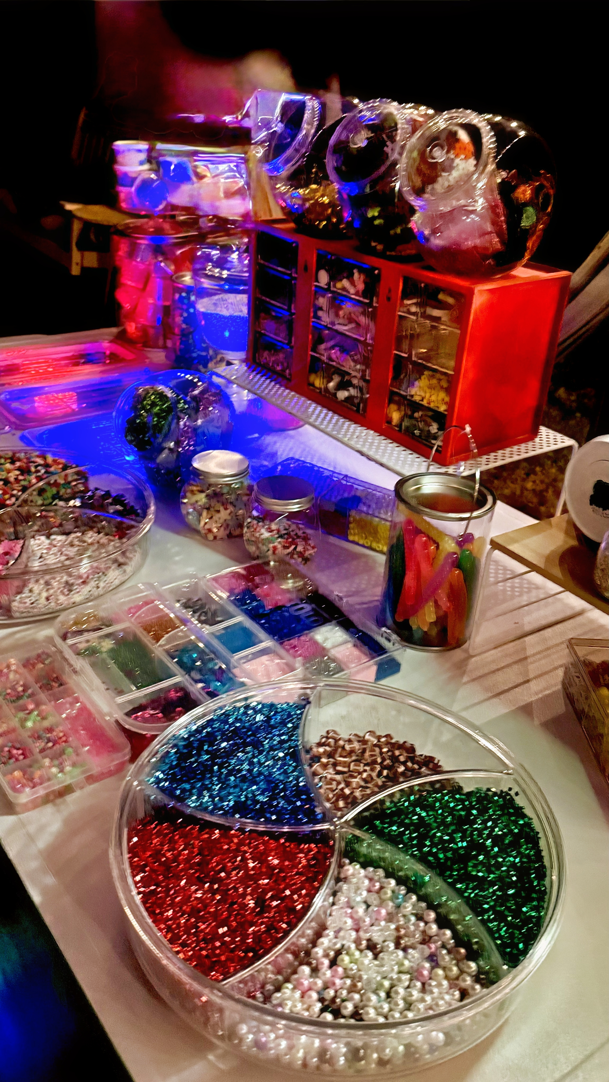 Candy display with various jars and containers of colorful candies, including gummy worms, jellies, sprinkles, and decorative beads.