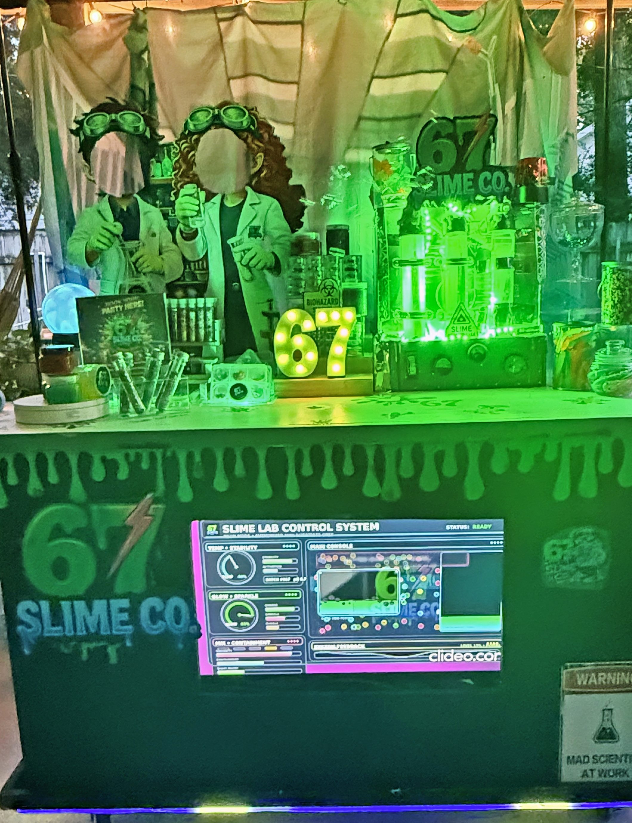 A colorful display featuring a sign for a slime lab control system, with various scientific equipment, containers, and decorations related to slime and science experiments. .  A visual of our 67 SLIME Co. Glow-In-The-Dark Slime Party.