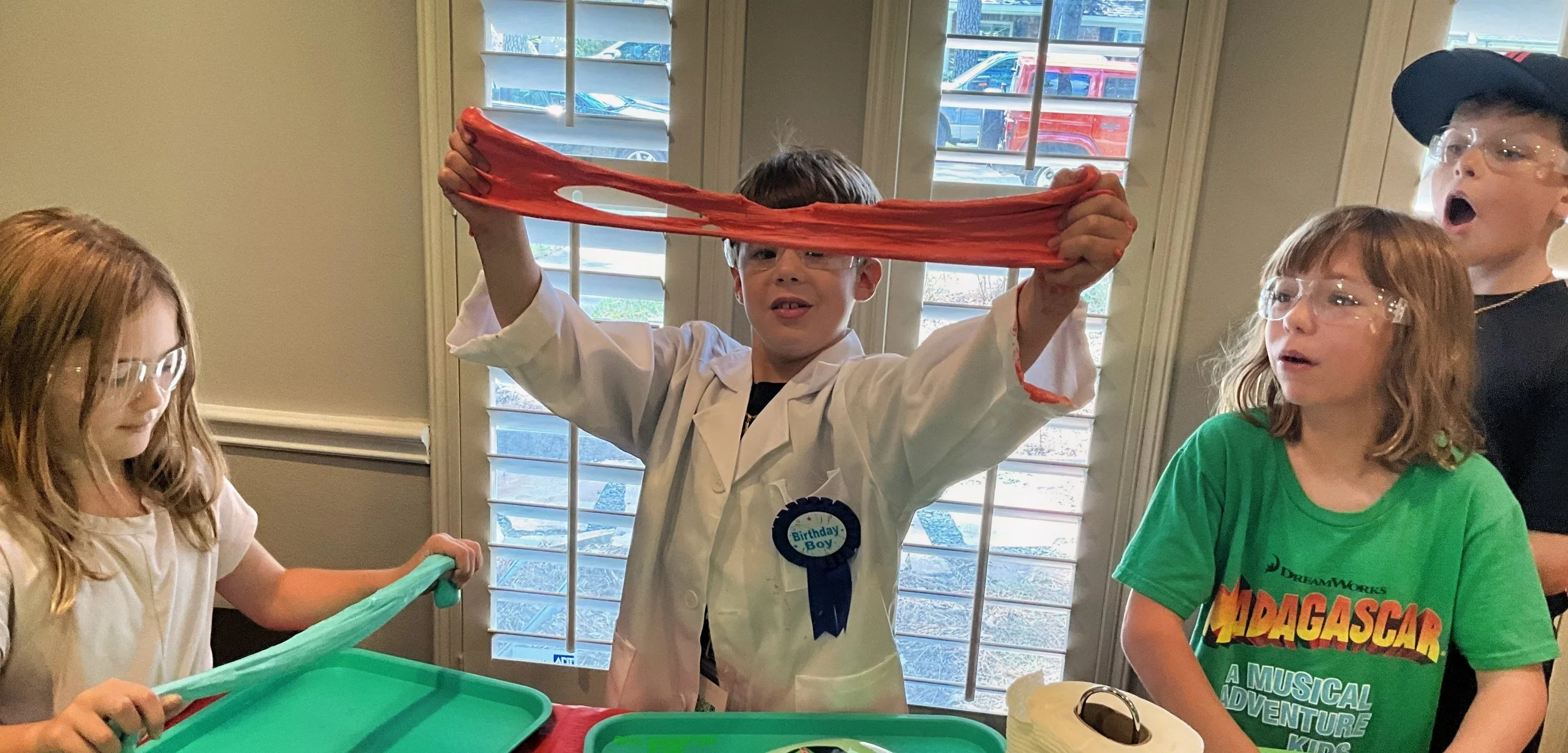 A boy is celebrating his birthday with friends & 67 SLIME Co. He is wearing a white lab coat and a badge that says "Birthday Boy." He is holding a large red slime.  They are all participating in a fun activity with slime.