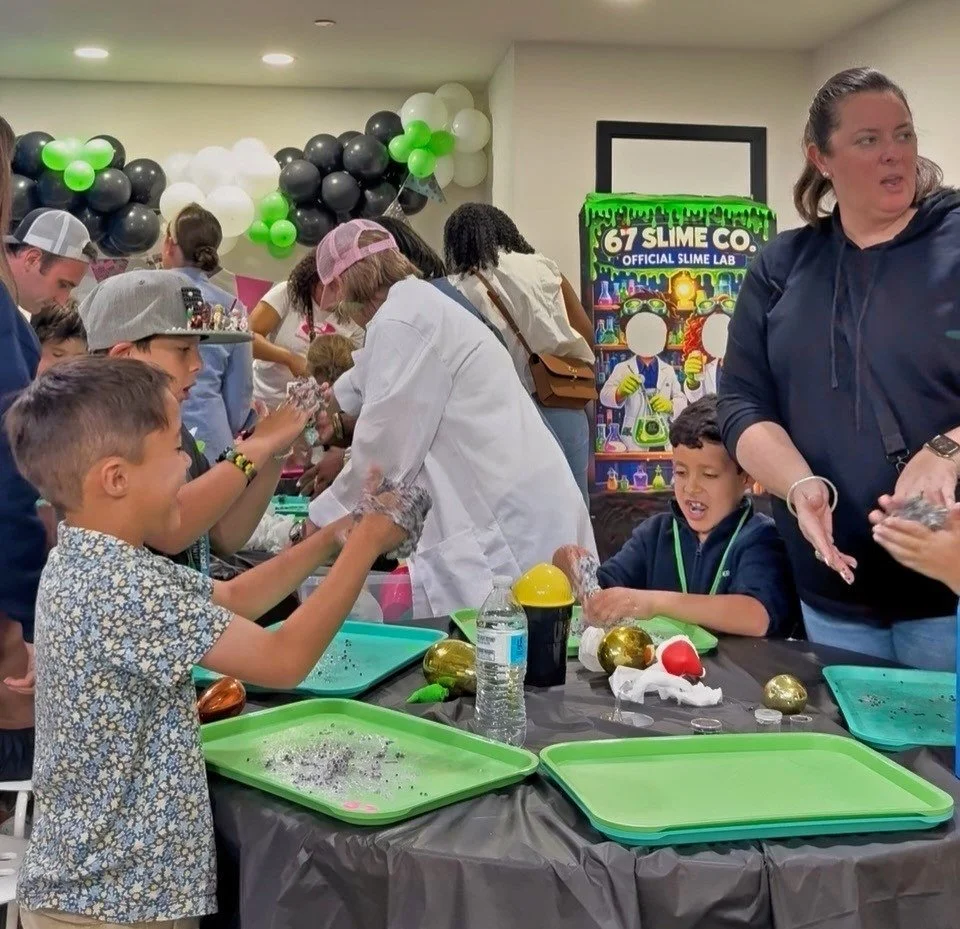 Children and adults participating in a slime-making activity at a birthday party or event, with balloons and a themed backdrop.