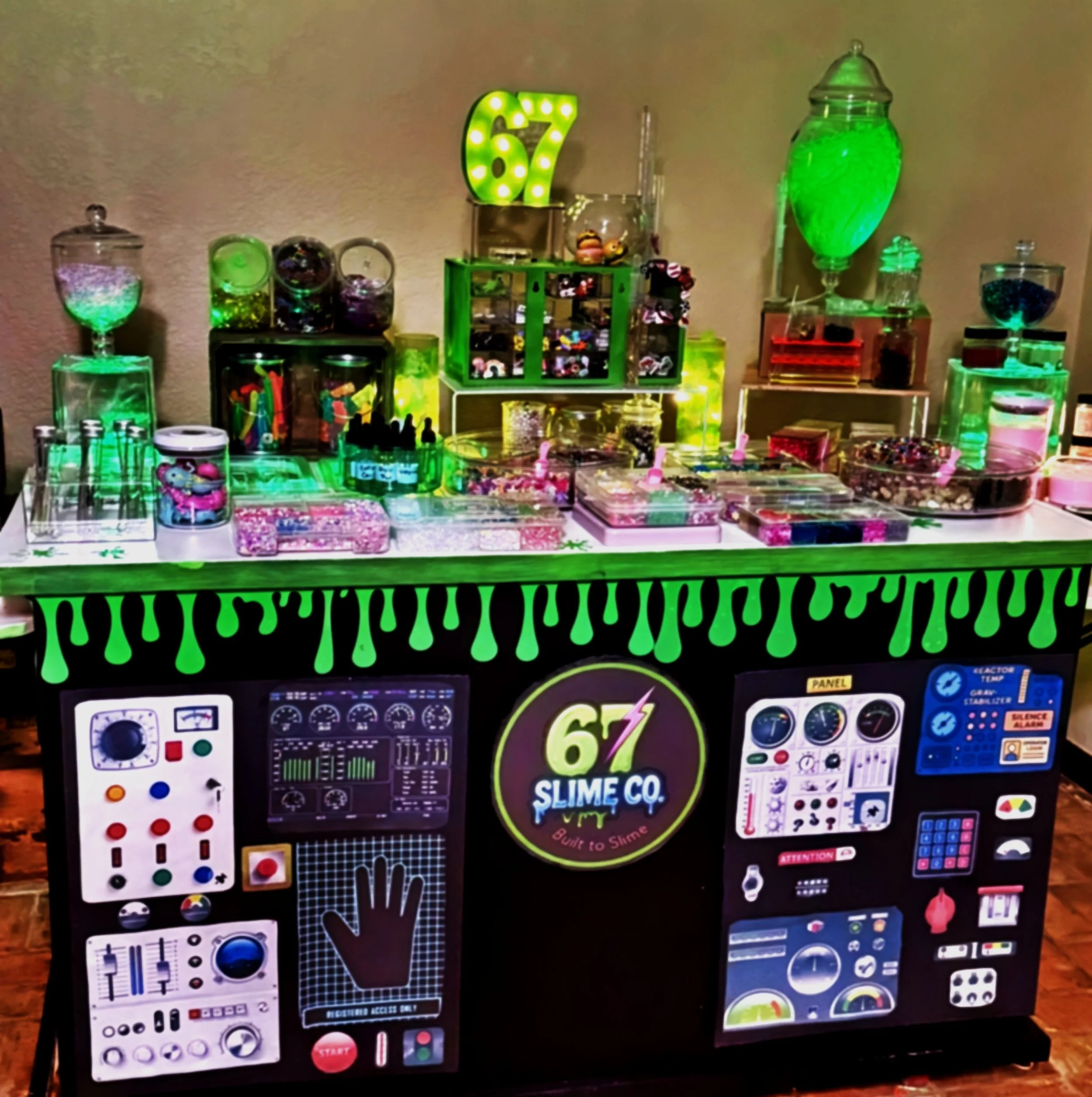 A colorful slime booth with various slime containers, tools, and decorations, featuring a brightly lit '67' sign and a chalkboard-style front panel with science-themed graphics and controls.