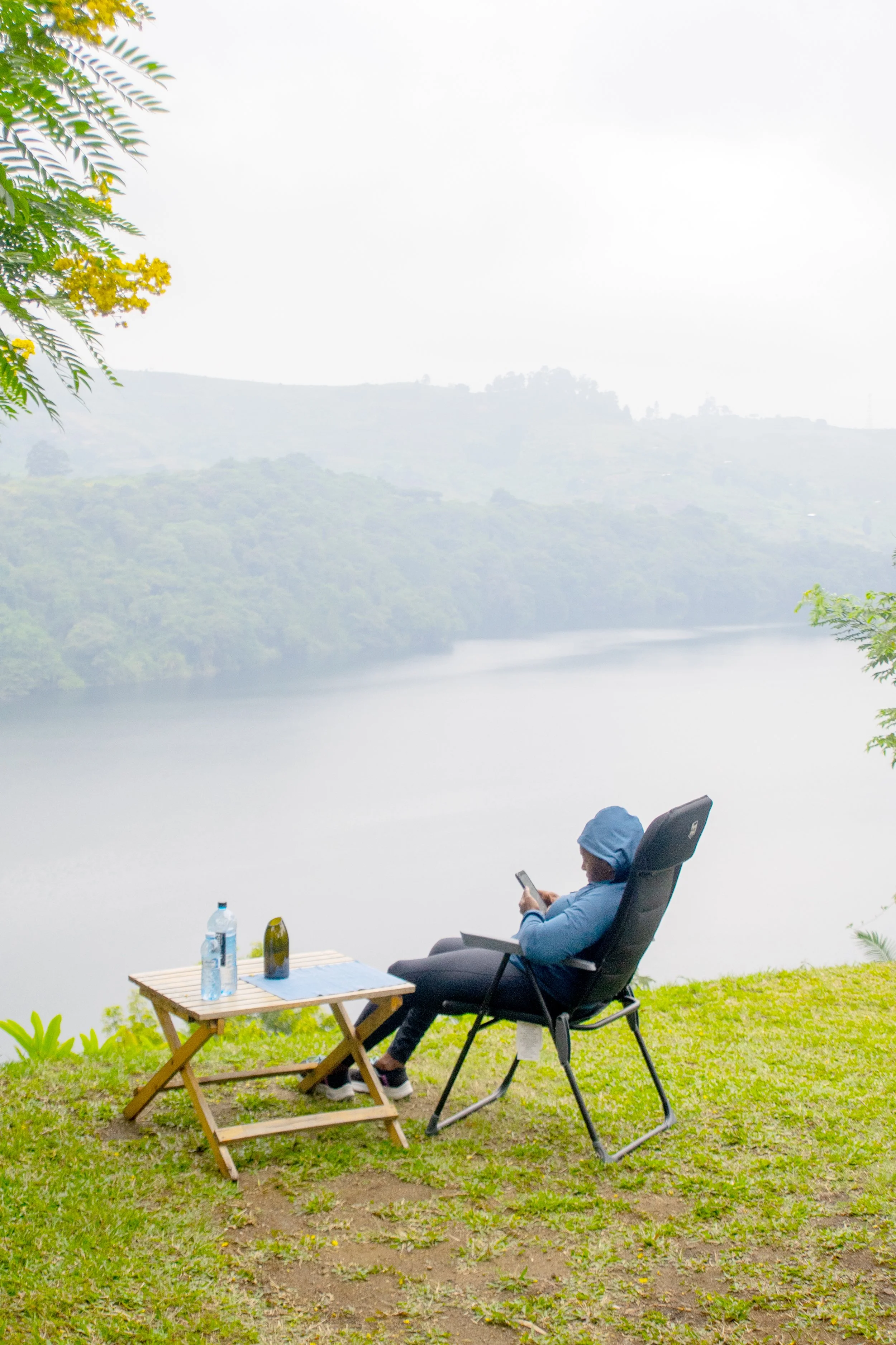 A person in blue clothing and a hood sitting in a black outdoor lounge chair on grassy ground near a lake or river, with trees and hills in the background, using a smartphone.
