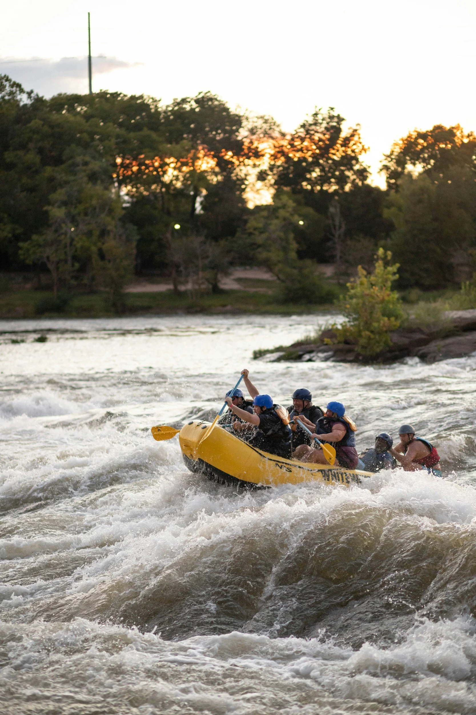 People white-water rafting during sunset on a river, wearing helmets and life jackets.