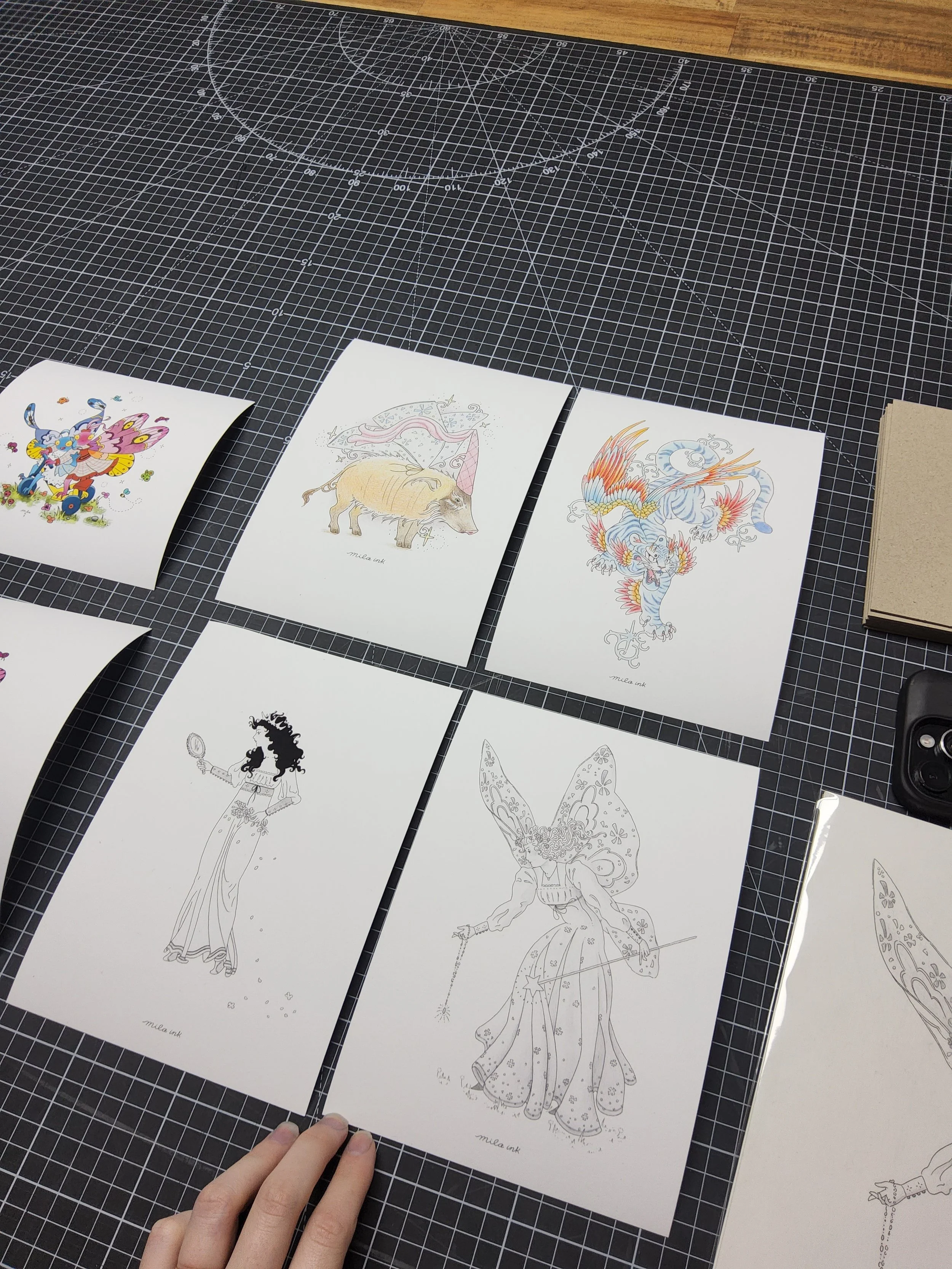 Sketches of colorful fantasy creatures and characters on white paper, placed on a black grid cutting mat, with a person's hand partially visible at the bottom.