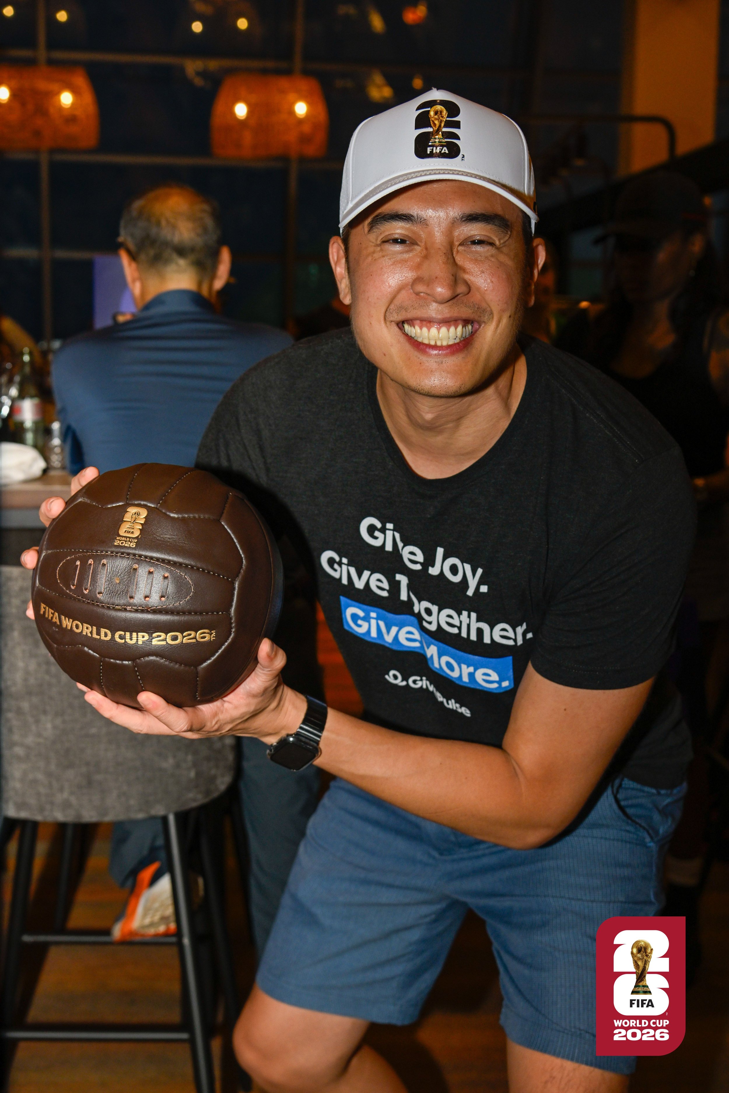 George Luc smiling and holding a brown FIFA World Cup 2026 soccer ball in a lively indoor setting with warm lighting and other people in the background.